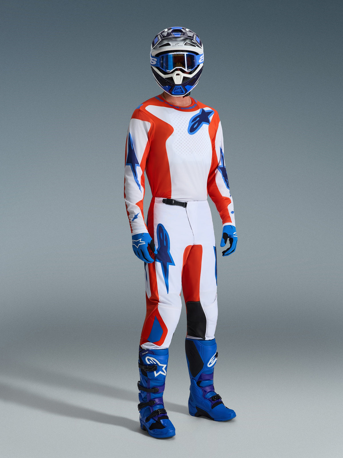 Piloto de motocross con el conjunto Alpinestars 2026 Fluid Grid, jersey y pantalones blancos con gráficos en naranja y azul UCLA, casco y gafas azules a juego, guantes azules y botas de competición en azul brillante, equipamiento de alto rendimiento coordinado para conducción off-road.