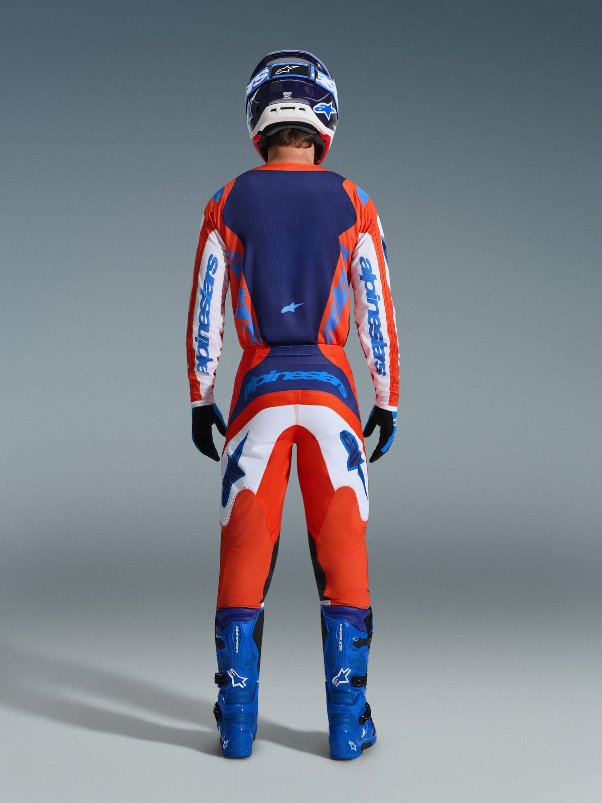 Alpinestars 2026 Fluid Grid Gear Set, piloto de motocross visto desde atrás con un conjunto completo de carreras en naranja y azul UCLA, jersey de manga larga con espalda azul marino y mangas blancas, pantalones naranjas a juego con gráficos azules, botas azules y casco coordinado, equipamiento profesional para carreras todoterreno.