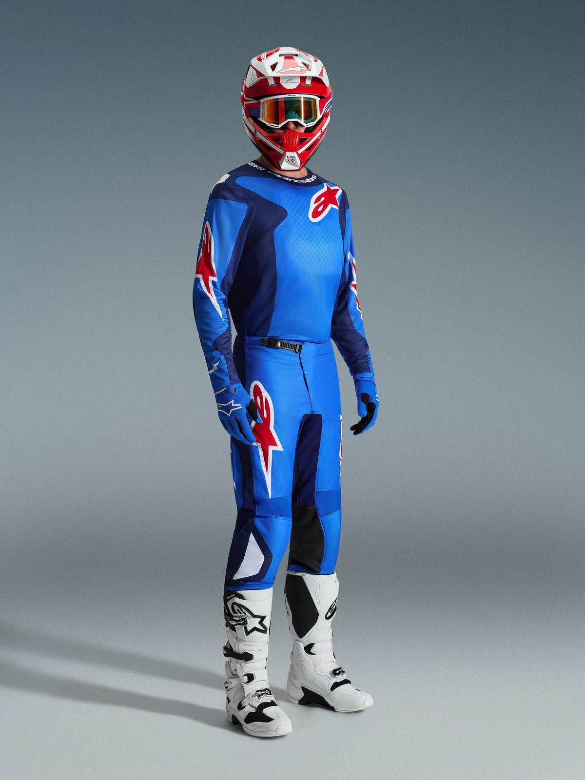 Piloto de motocross con el Jersey Alpinestars Fluid Grid 2026 en azul y rojo, combinado con pantalones de motocross a juego, botas blancas y negras y un casco rojo y blanco, conjunto de equipamiento profesional para rendimiento off-road.