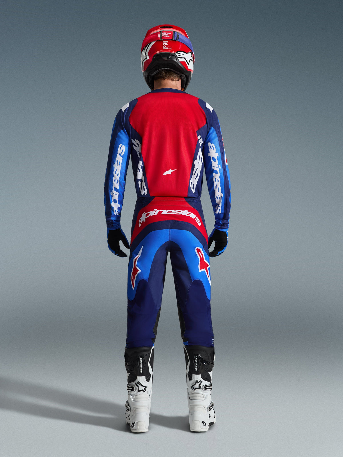 Vista posterior de un piloto de motocross con el Jersey Fluid Grid 2026 en azul y rojo, con un gran panel trasero rojo y mangas azules con la marca Alpinestars en blanco, combinado con pantalones a juego, botas blancas y negras, y un casco rojo para un equipo completo de motocross.