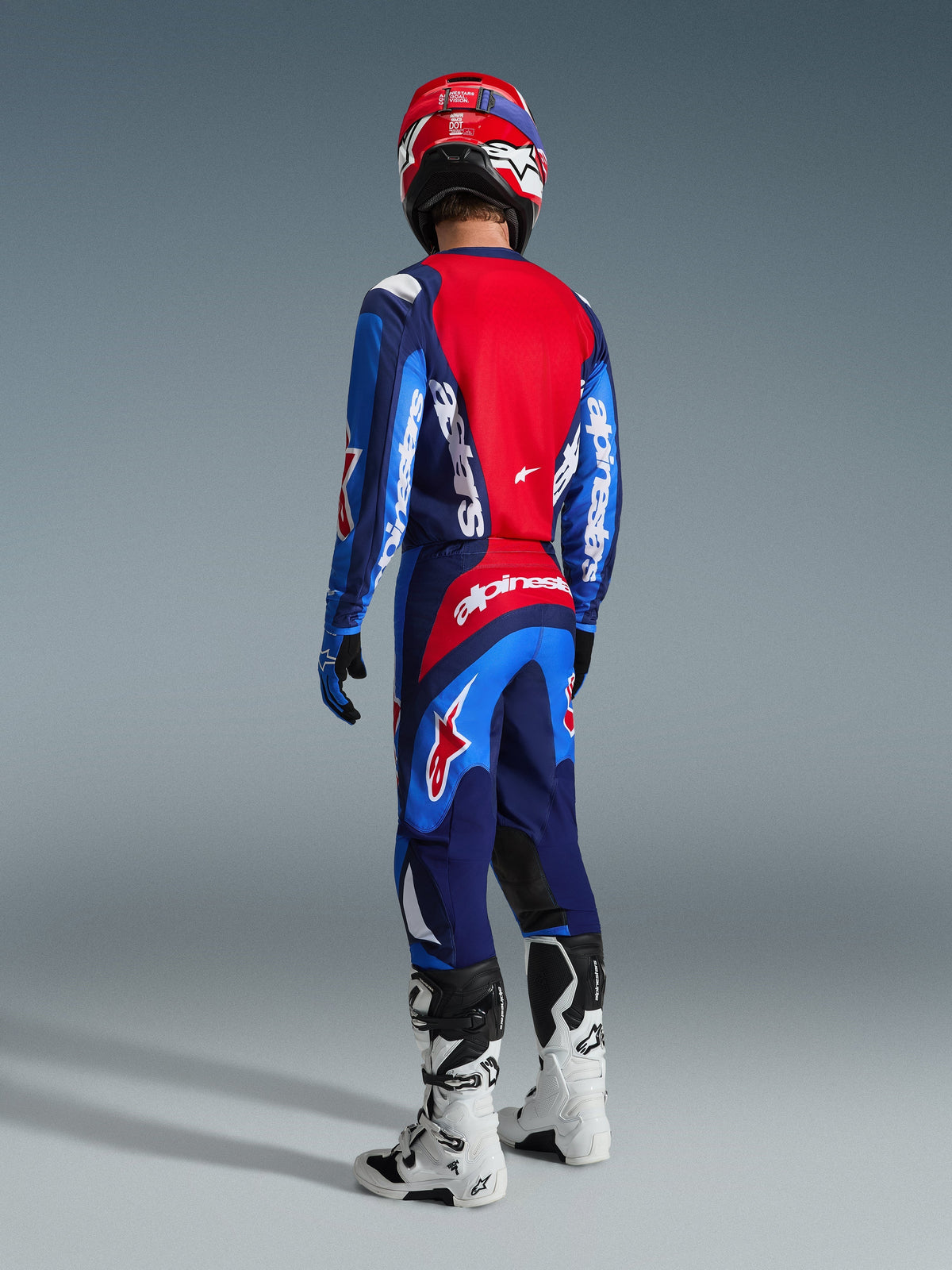 Piloto de motocross desde la parte posterior vistiendo el jersey Alpinestars 2026 Fluid Grid en azul y rojo, pantalones de motocross a juego con logotipos blancos llamativos, casco rojo y botas blancas y negras, conjunto completo de equipamiento para carreras off-road