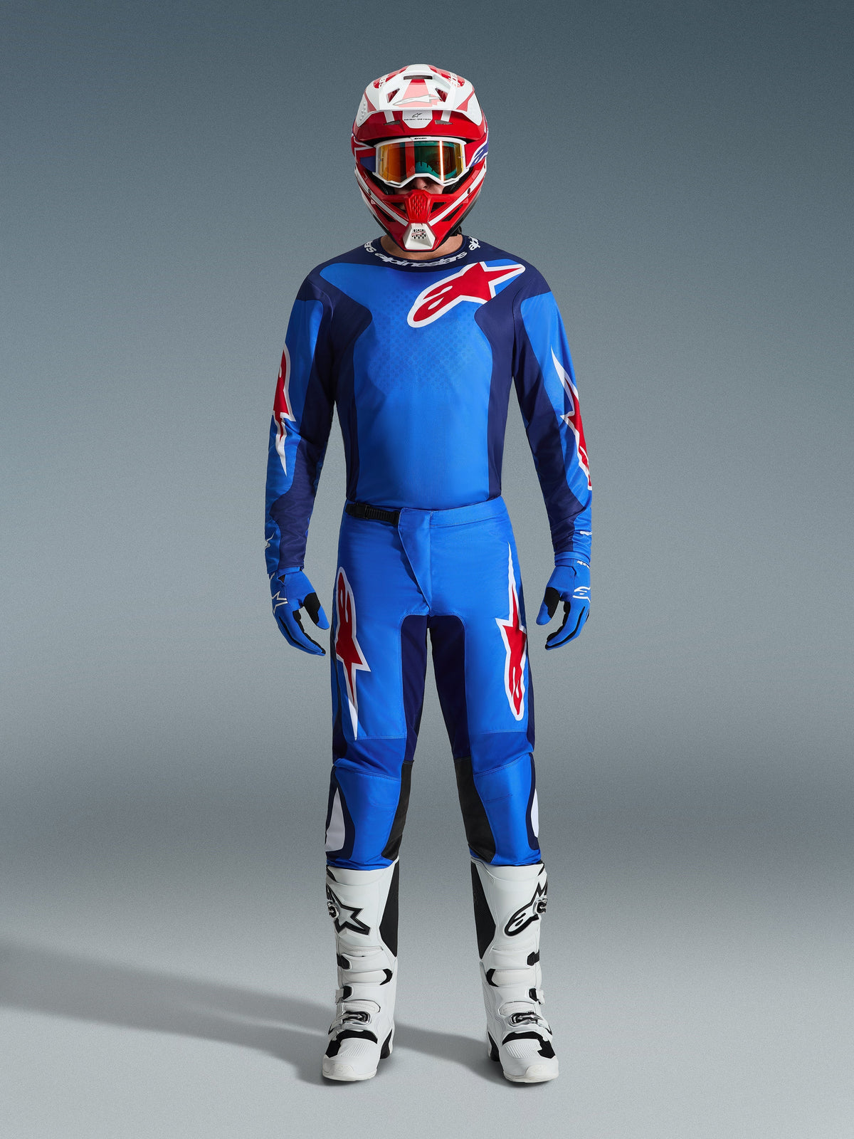 Piloto de motocross con el Jersey Alpinestars Fluid Grid 2026 en azul y rojo, pantalones azules a juego con logotipos de estrellas blancas y rojas, casco rojo y blanco con gafas de espejo, guantes azules y botas de carreras blancas y negras, conjunto completo de equipamiento para todoterreno.