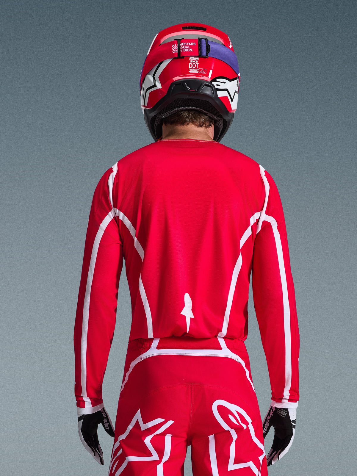Vista posterior de un piloto de motocross que viste el Jersey Alpinestars Fluid Apex 2026 en rojo y blanco, con patrones geométricos blancos, combinado con pantalones de motocross a juego y un casco rojo, equipamiento de competición completo para rendimiento off-road