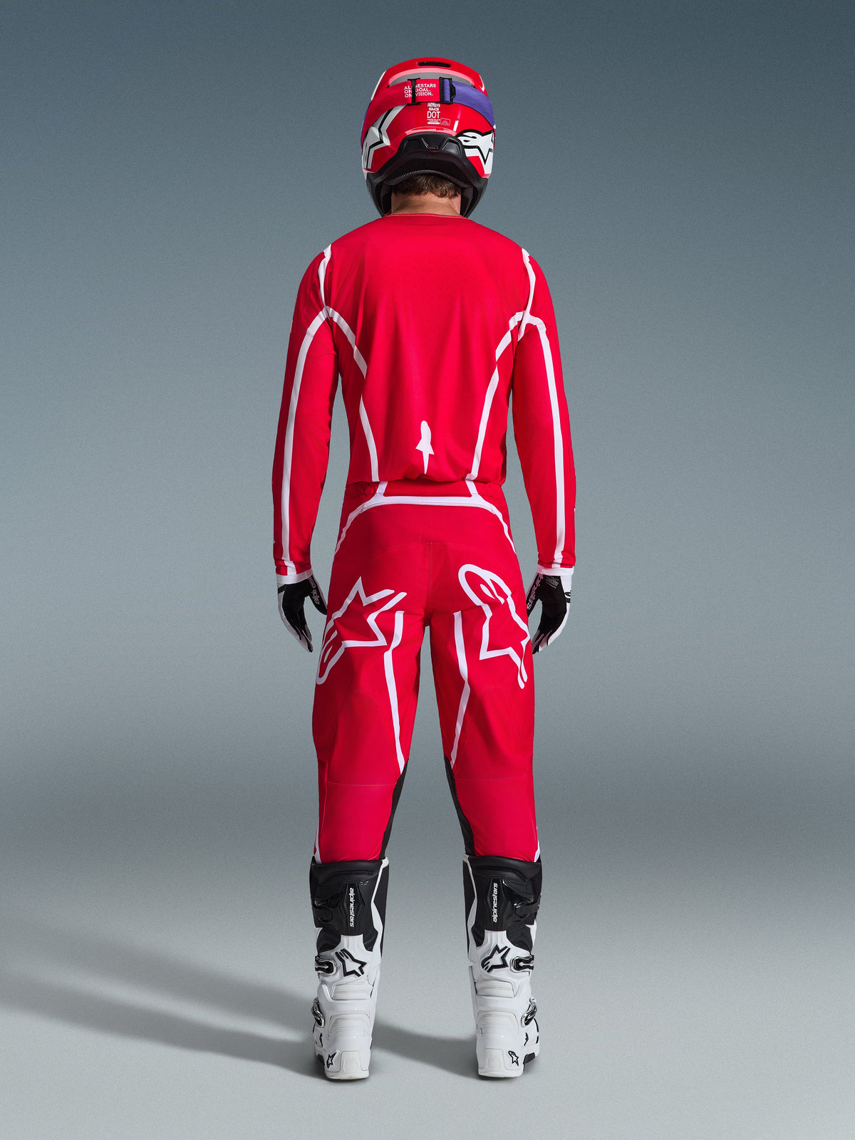 Vista trasera de un piloto de motocross con el conjunto Alpinestars 2026 Fluid Apex, jersey y pantalones de color rojo brillante con franjas y logotipos blancos, combinados con botas blancas y negras y un casco rojo, equipamiento profesional de competición off-road
