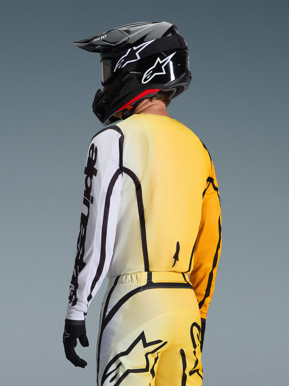 Vista trasera de tres cuartos de un piloto de motocross que viste el Jersey Fluid Apex 2026 en Yellow Black, con un diseño degradado de amarillo a blanco, marca Alpinestars en negro en la manga, usado con pantalones a juego y un casco de motocross negro.