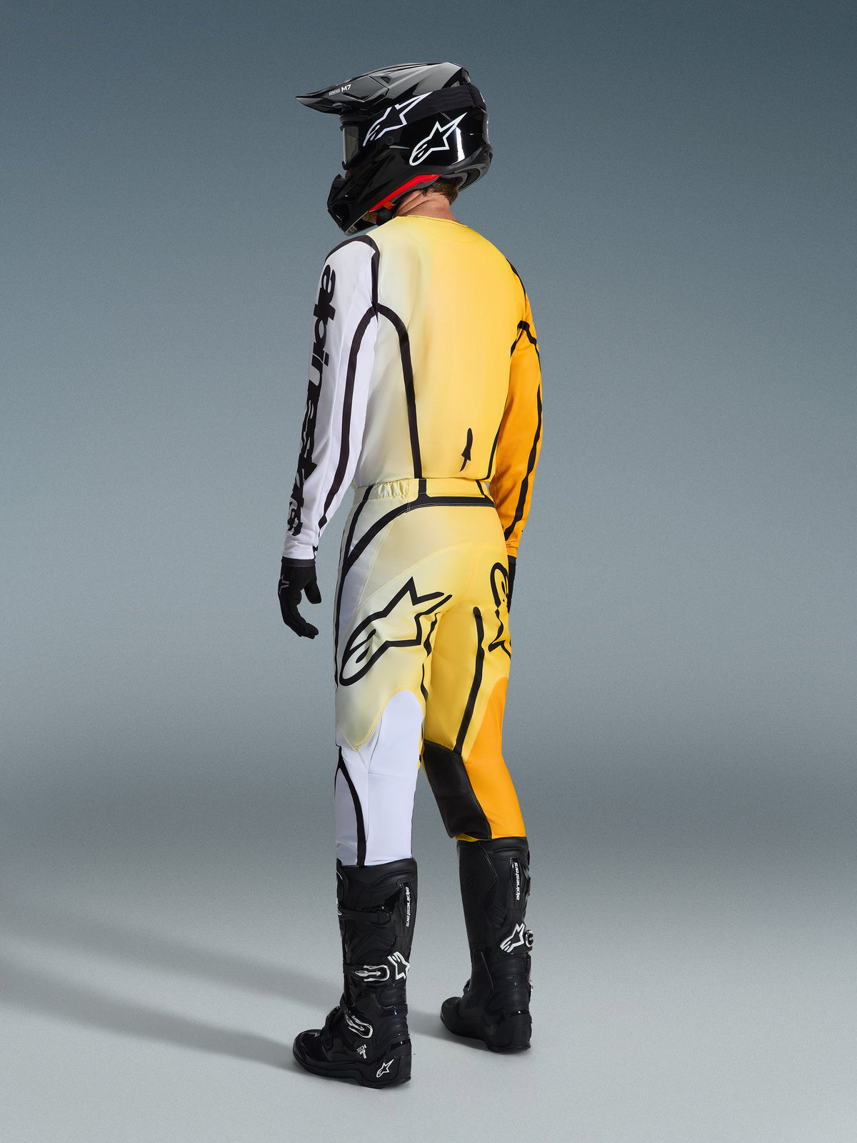 Piloto de motocross visto desde atrás vistiendo el Jersey Alpinestars Fluid Apex 2026 y pantalones a juego, conjunto amarillo y negro con degradados de amarillo a blanco y líneas gráficas negras marcadas, combinado con casco negro, guantes blancos y negros, y botas de motocross negras