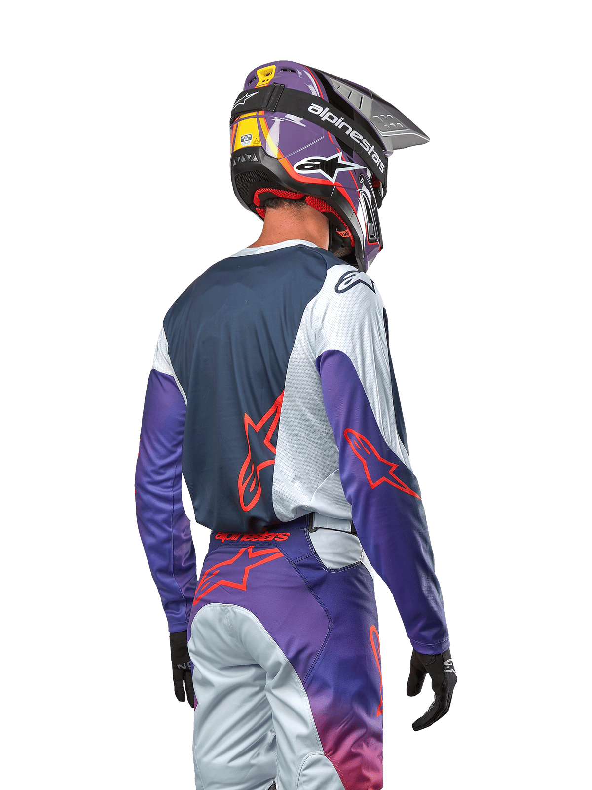 Piloto de motocross visto desde atrás con el Jersey Racer Hoen 2024, panel trasero azul marino con hombros gris claro y mangas moradas, logotipos Alpinestars en naranja intenso, pantalones a juego en morado y gris con casco de motocross morado, equipamiento completo de carreras off-road