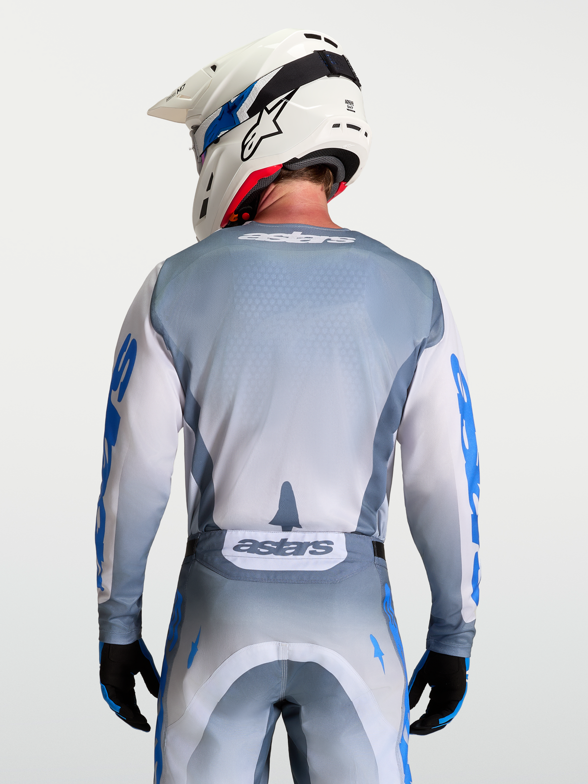 Alpinestars Jersey Racer Amber, jersey de motocross gris y blanco para hombre, vista trasera en modelo con casco blanco, materiales técnicos transpirables y gráficos en azul