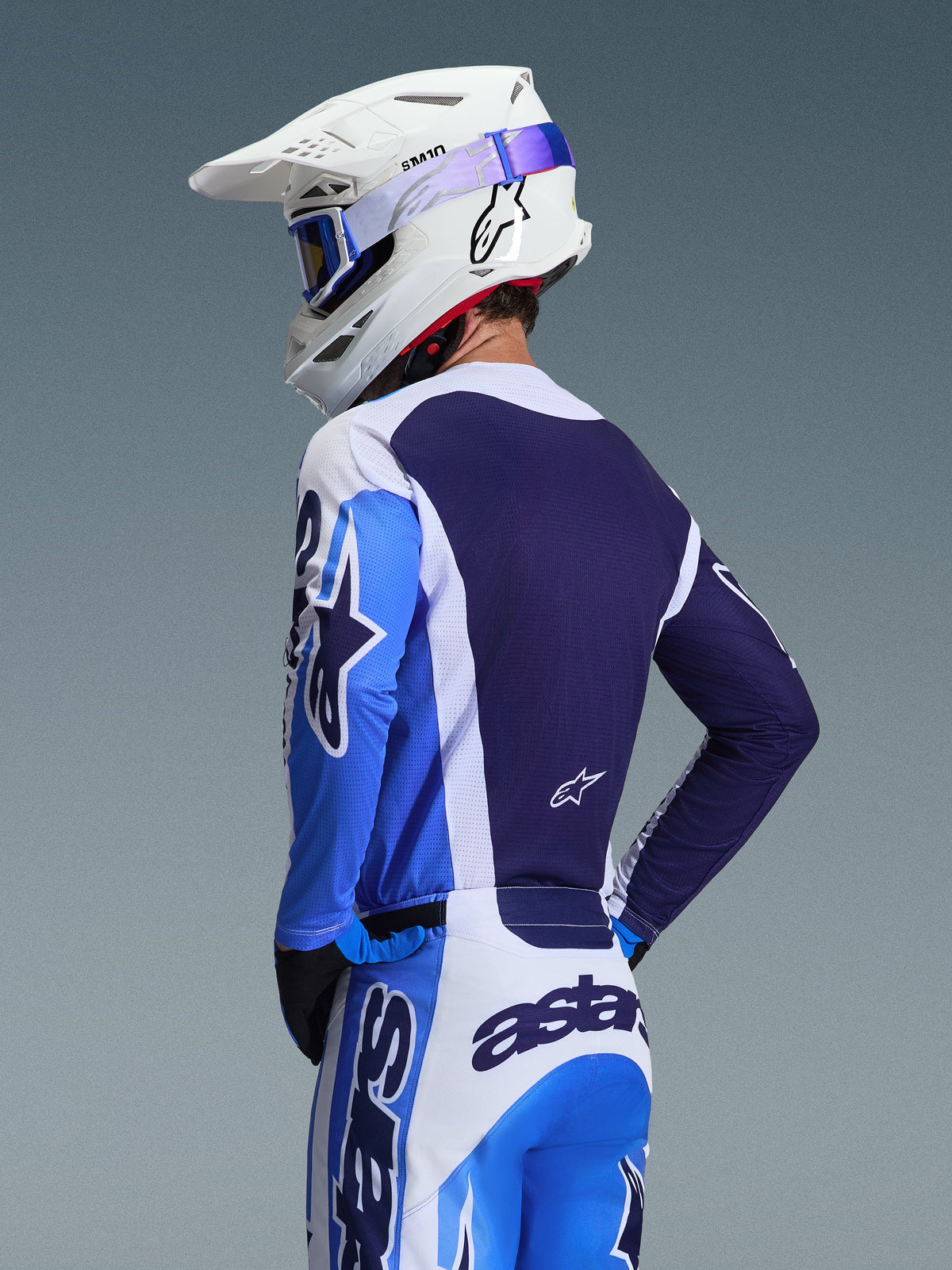Piloto de motocross luciendo el jersey Alpinestars 2026 Racer Air Portl en azul Ucla y blanco, vista posterior que destaca el panel trasero de malla transpirable en azul marino, con pantalones a juego, casco Alpinestars blanco y gafas azules, conjunto de equipamiento de motocross coordinado.
