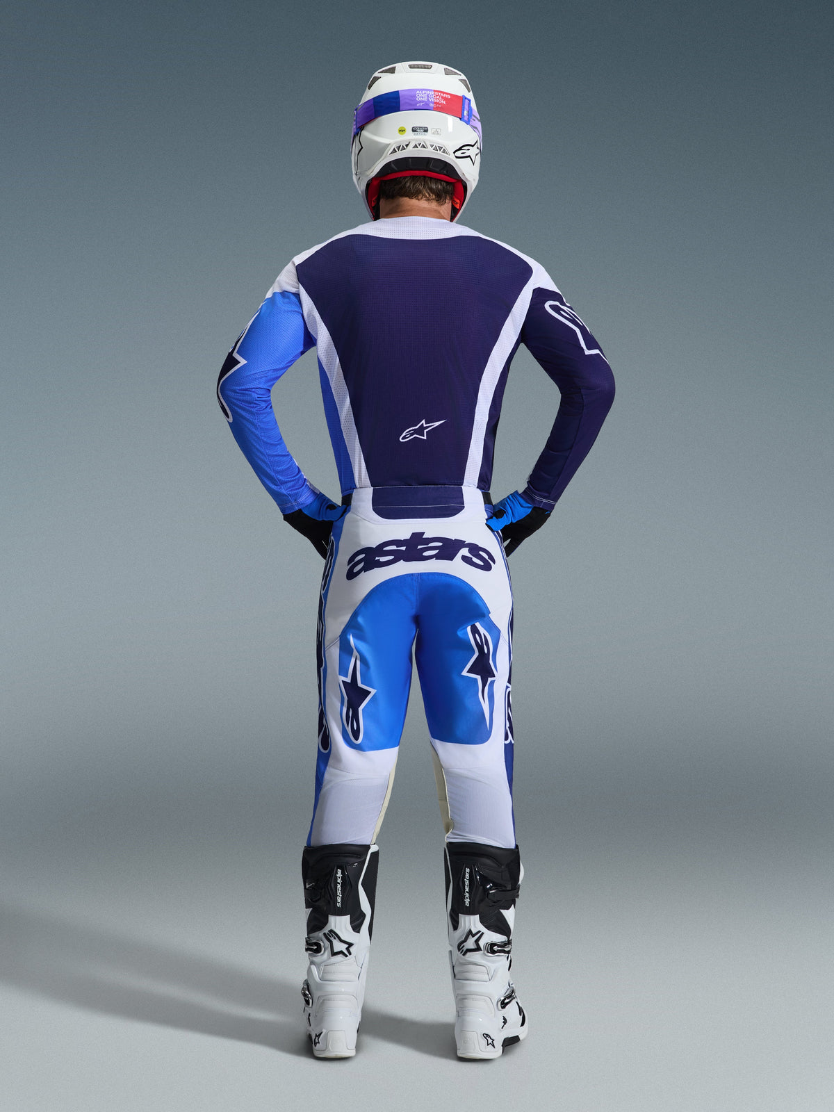 Vista posterior de un piloto de motocross con el Jersey Alpinestars Racer Air Portl 2026 en color Ucla Blue White, combinado con pantalones blancos y azules a juego y botas blancas, destacando la construcción de malla de flujo de aire completo en la espalda para carreras off-road.