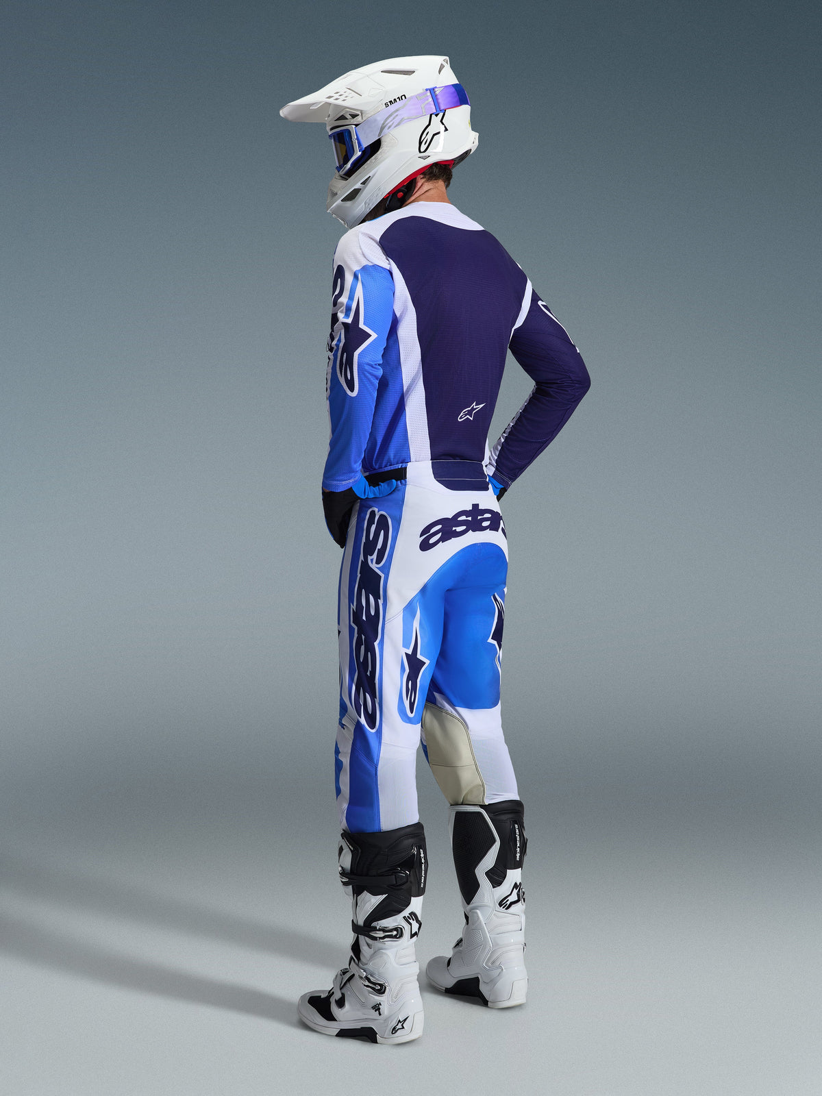 Piloto de motocross desde una perspectiva trasera vistiendo el Jersey Alpinestars 2026 Racer Air Portl en color Ucla Blue y blanco, combinado con pantalones de motocross a juego, casco Supertech blanco y botas blancas, mostrando un conjunto de equipamiento de competición coordinado.
