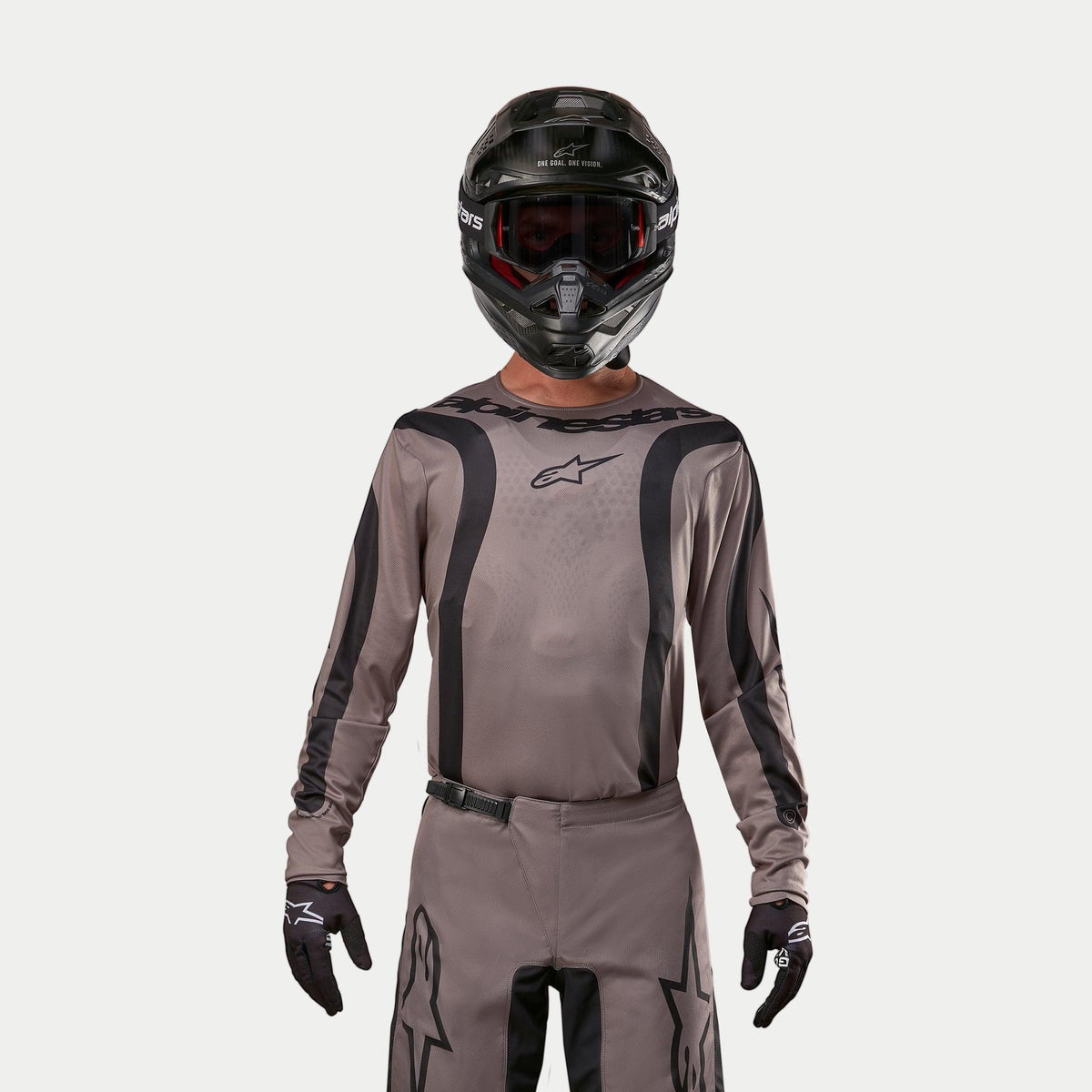 Piloto de motocross con el Jersey Alpinestars 2024 Fluid Lurv en color Mud Black, un tejido gris terroso con franjas gráficas negras y un logo de estrella en el pecho, combinado con casco negro, gafas rojas y guantes negros para un look de carreras todoterreno coordinado.