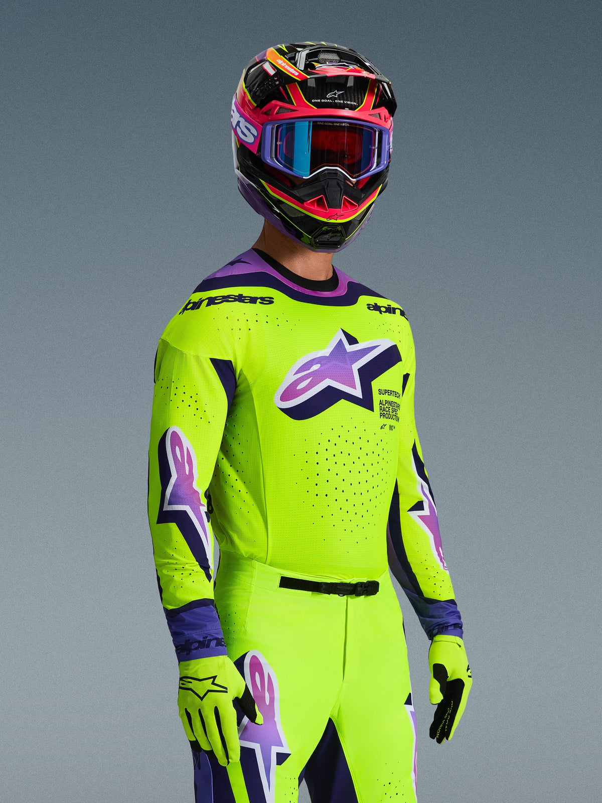 Piloto de motocross con el jersey Alpinestars 2026 Supertech Vista y conjunto de equipamiento a juego, amarillo brillante con detalles en púrpura y negro, con un gran logotipo Astar púrpura en el pecho y las mangas, guantes a juego y casco de competición multicolor con gafas, ajuste atlético de nivel profesional para carreras off-road.