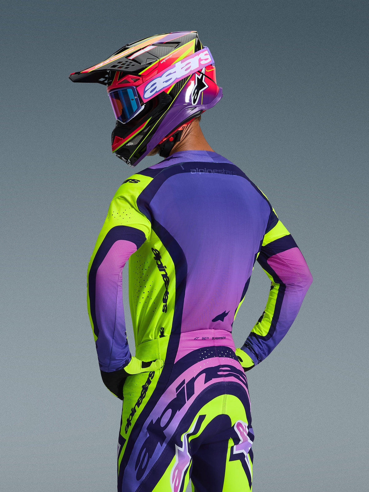 Piloto de motocross visto desde atrás con el conjunto Alpinestars Supertech Vista 2026, jersey y pantalones en amarillo, púrpura y negro, con un diseño de degradado vibrante, ventilación de malla y logotipos llamativos de Alpinestars, combinado con un casco y gafas púrpura.