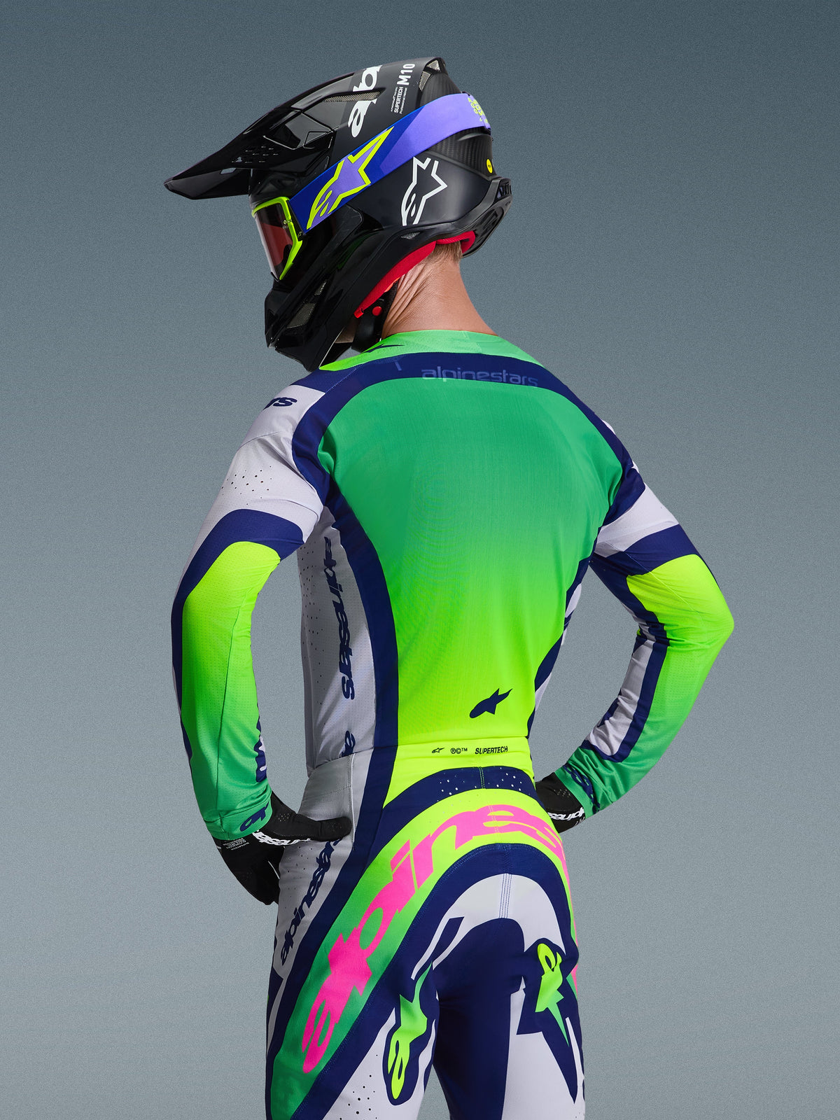 Vista trasera de un piloto de motocross con el conjunto Alpinestars 2026 Supertech Vista, que incluye un jersey verde y blanco con detalles en azul marino, pantalones a juego con un destacado logotipo rosa de Alpinestars, casco de competición negro con gráficos azules y gafas de lente roja sobre un fondo gris neutro.