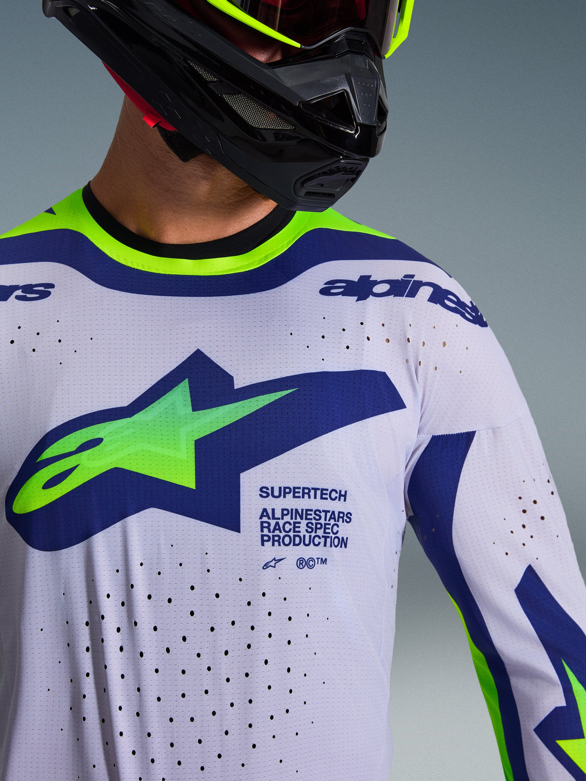 Primer plano del Jersey Alpinestars Supertech Vista 2026 en gris claro, amarillo flúor y azul, usado por un piloto con casco negro, con un gran logotipo en el pecho, perforaciones de ventilación cortadas con láser y gráficos de competición de nivel profesional.