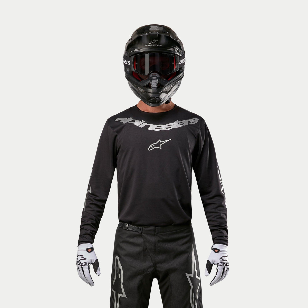 Jersey Alpinestars Fluid Graphite 2024, jersey de motocross negro y plateado, usado por un piloto con casco negro y gafas rojas, con la marca Alpinestars en plata en el pecho, combinado con pantalones negros y guantes blancos para un rendimiento off-road.