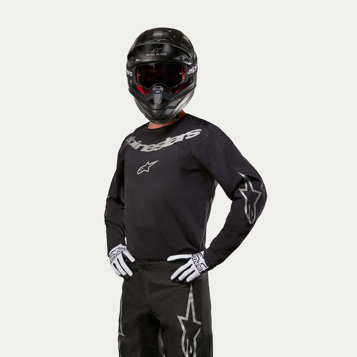 Piloto de motocross con el Jersey Alpinestars Fluid Graphite 2024 en color negro y plateado, combinado con pantalones de motocross negros, casco negro con gafas de lentes rojas y guantes blancos y negros, con logotipos plateados de la marca en el pecho y las mangas.