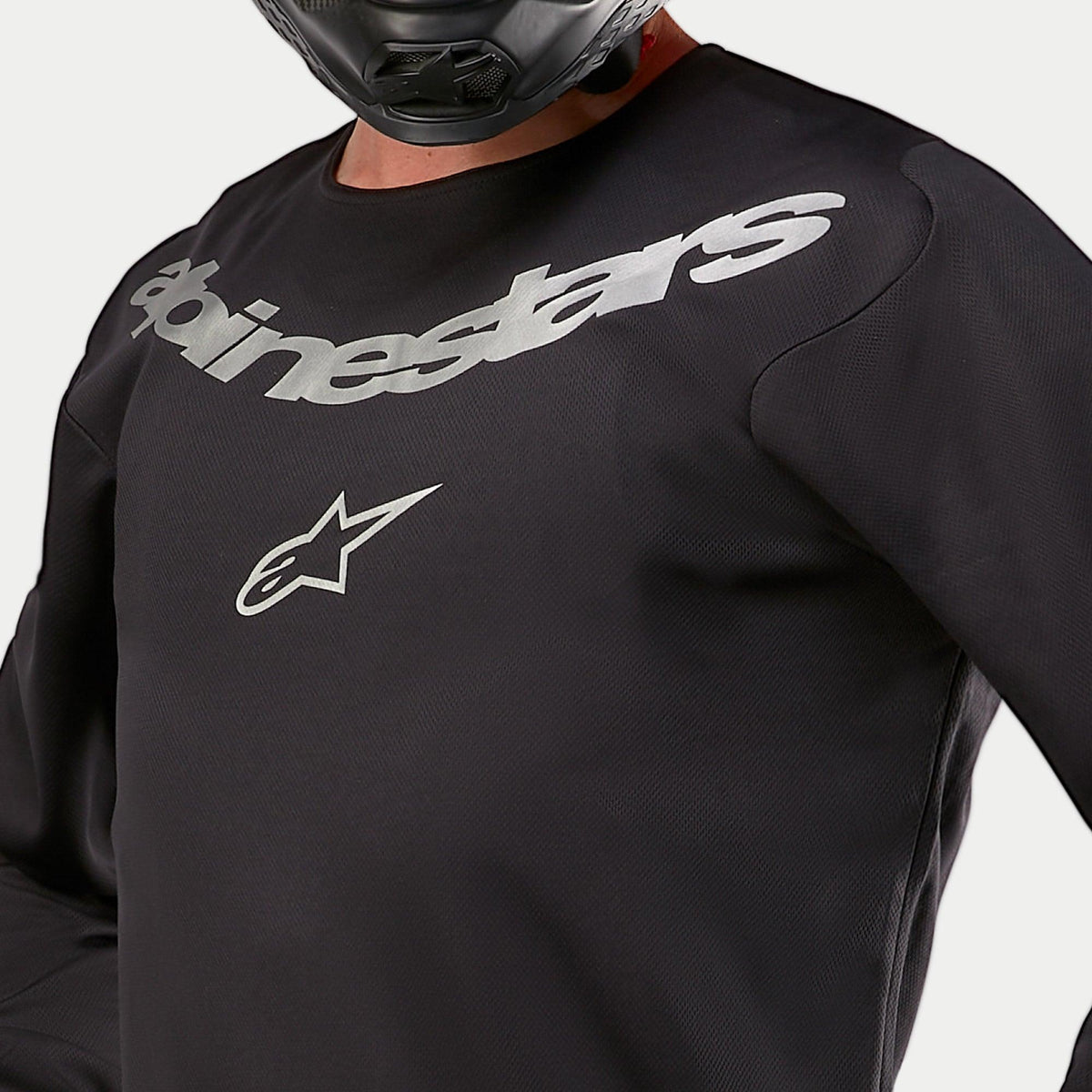 Jersey Alpinestars Fluid Graphite 2024, jersey de motocross, negro y plata, detalle en primer plano del área del pecho con la marca y el logotipo de la estrella de Alpinestars en plata, construcción de poli-tejido transpirable, usado por un piloto con casco negro