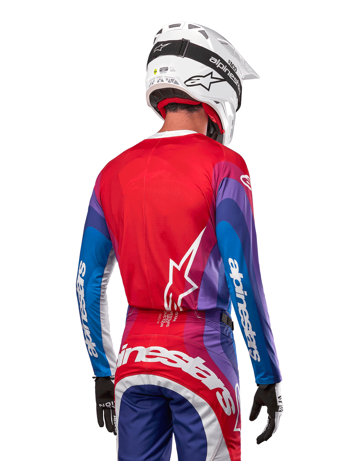 Piloto de motocross visto desde atrás luciendo el Jersey Alpinestars Techstar Pneuma 2024 en azul, rojo marte y blanco, con espalda roja y logo de estrella blanco, mangas azules y moradas con marca en blanco, combinado con pantalones a juego y casco blanco para carreras off-road.