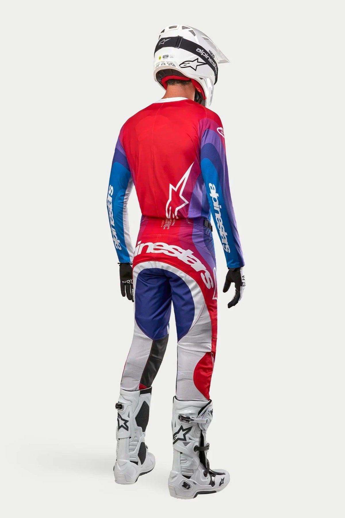Motocross rider from behind wearing Alpinestars 2024 Techstar Pneuma jersey and pants, Blu Mars Red White colorway with red, blue, and purple gradients, featuring white branding, paired with white boots and a white helmet, coordinated off-road racing gear.