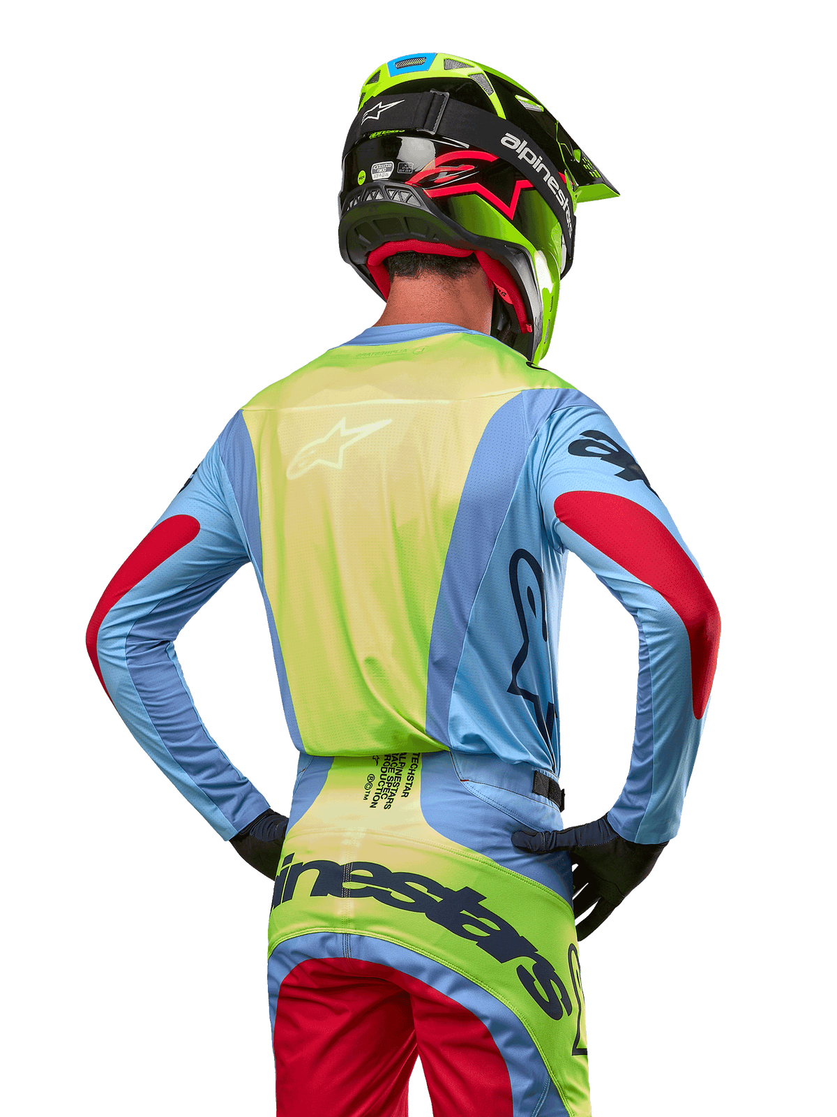 Piloto de motocross desde atrás vistiendo el Jersey Alpinestars Techstar Ocuri 2024, azul claro con espalda en amarillo flúor y detalles rojos en las mangas, pantalones de motocross a juego y casco amarillo neón, conjunto coordinado para carreras fuera de carretera