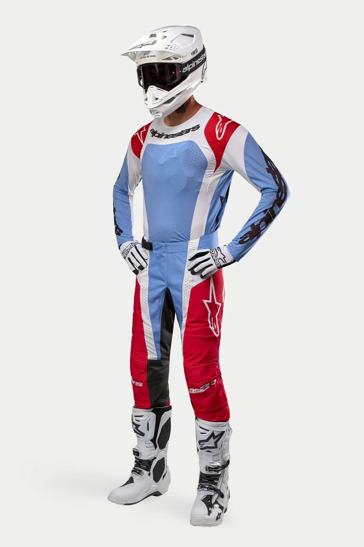 Piloto de motocross con equipamiento completo de carreras, incluyendo el jersey Alpinestars 2024 Techstar Ocuri en azul claro, rojo Marte y blanco, pantalones a juego, casco blanco con gafas, botas blancas y guantes blancos y negros.
