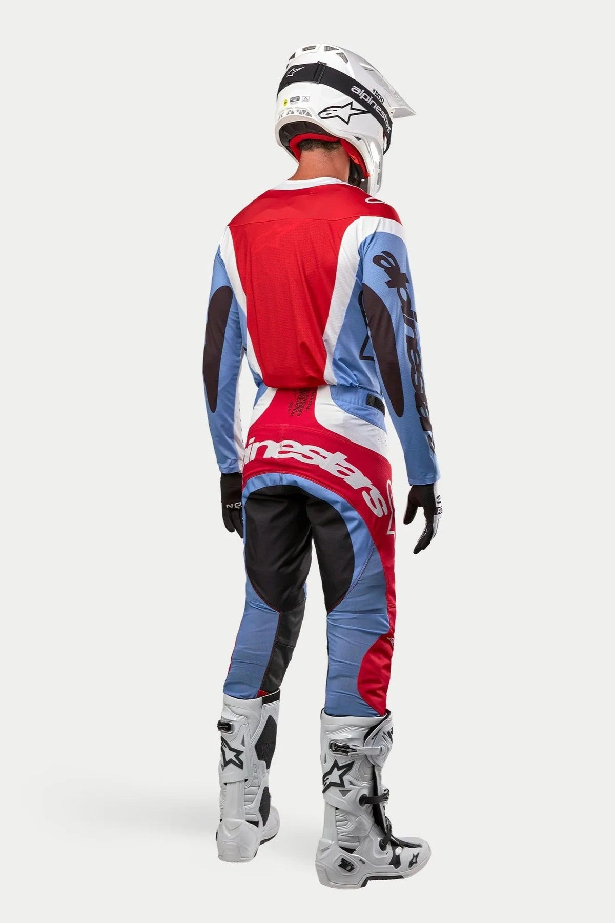 Piloto de motocross visto desde atrás con equipamiento de competición completo, Jersey Techstar Ocuri 2024 en azul claro, rojo Marte y blanco, pantalones a juego con marca llamativa, botas blancas y negras, casco blanco, conjunto coordinado para carreras todoterreno