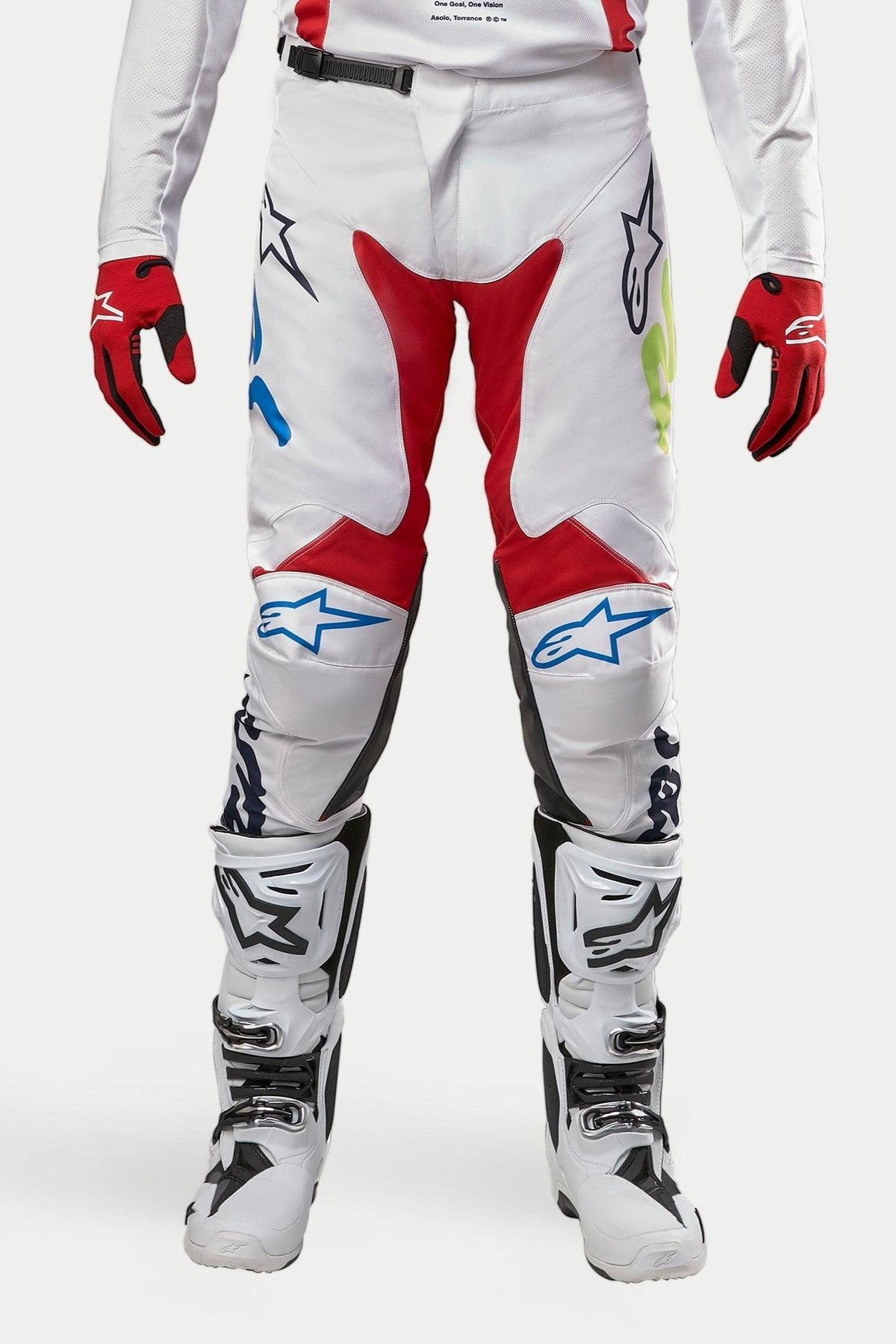 Alpinestars 2024 Racer Hana Pants, pantalones de motocross, blanco multicolor con detalles en rojo, azul y lima, usados con botas blancas y negras y guantes rojos, con logotipos de Alpinestars en las rodillas y un diseño técnico duradero para carreras todoterreno
