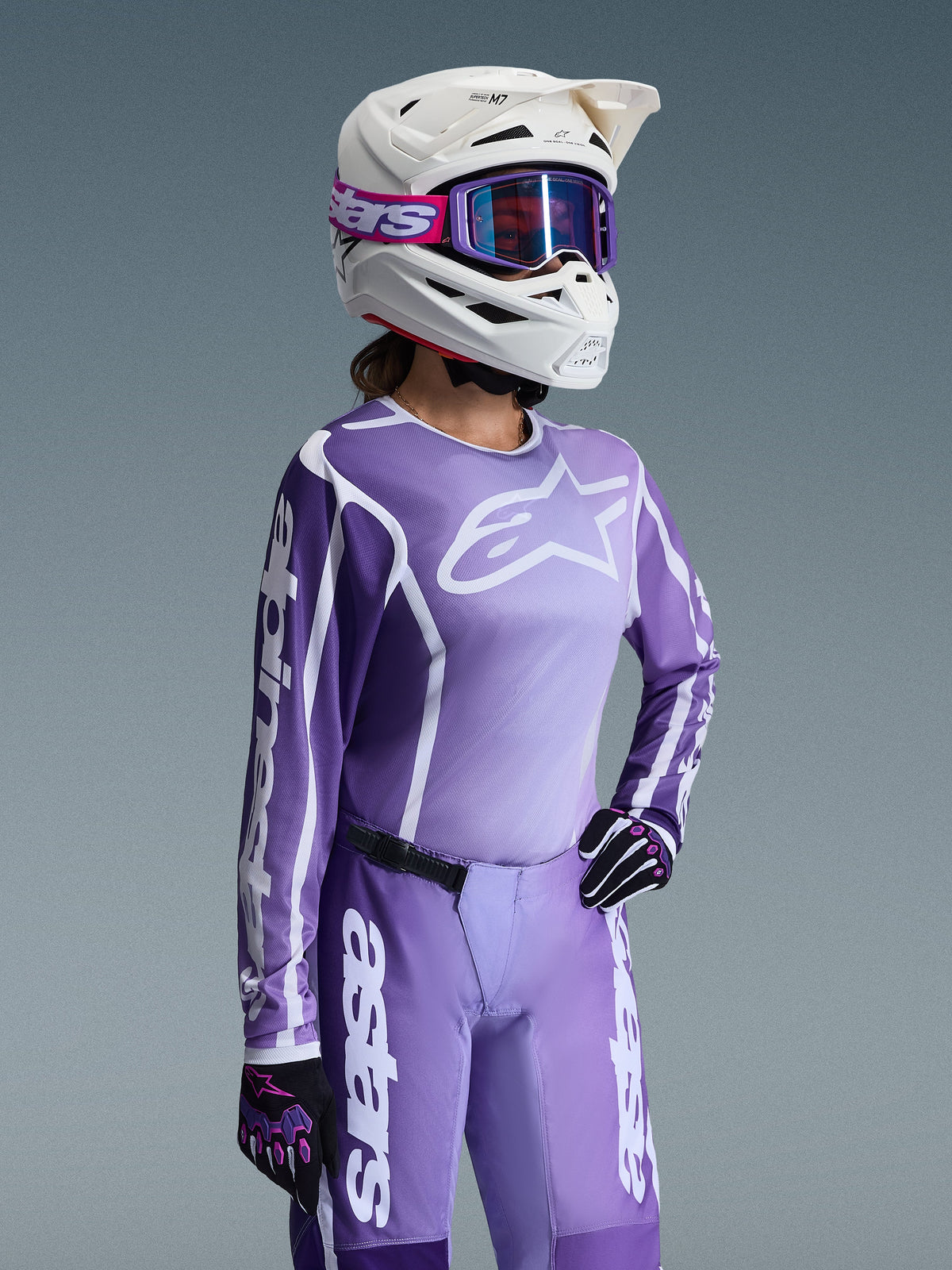 Piloto de motocross con el conjunto de equipamiento Alpinestars Stella Fluid Apex 2026 para mujer, jersey morado y blanco con logotipos de Alpinestars, pantalones morados a juego, casco blanco con gafas moradas y guantes negros y morados, equipo profesional de carreras todoterreno