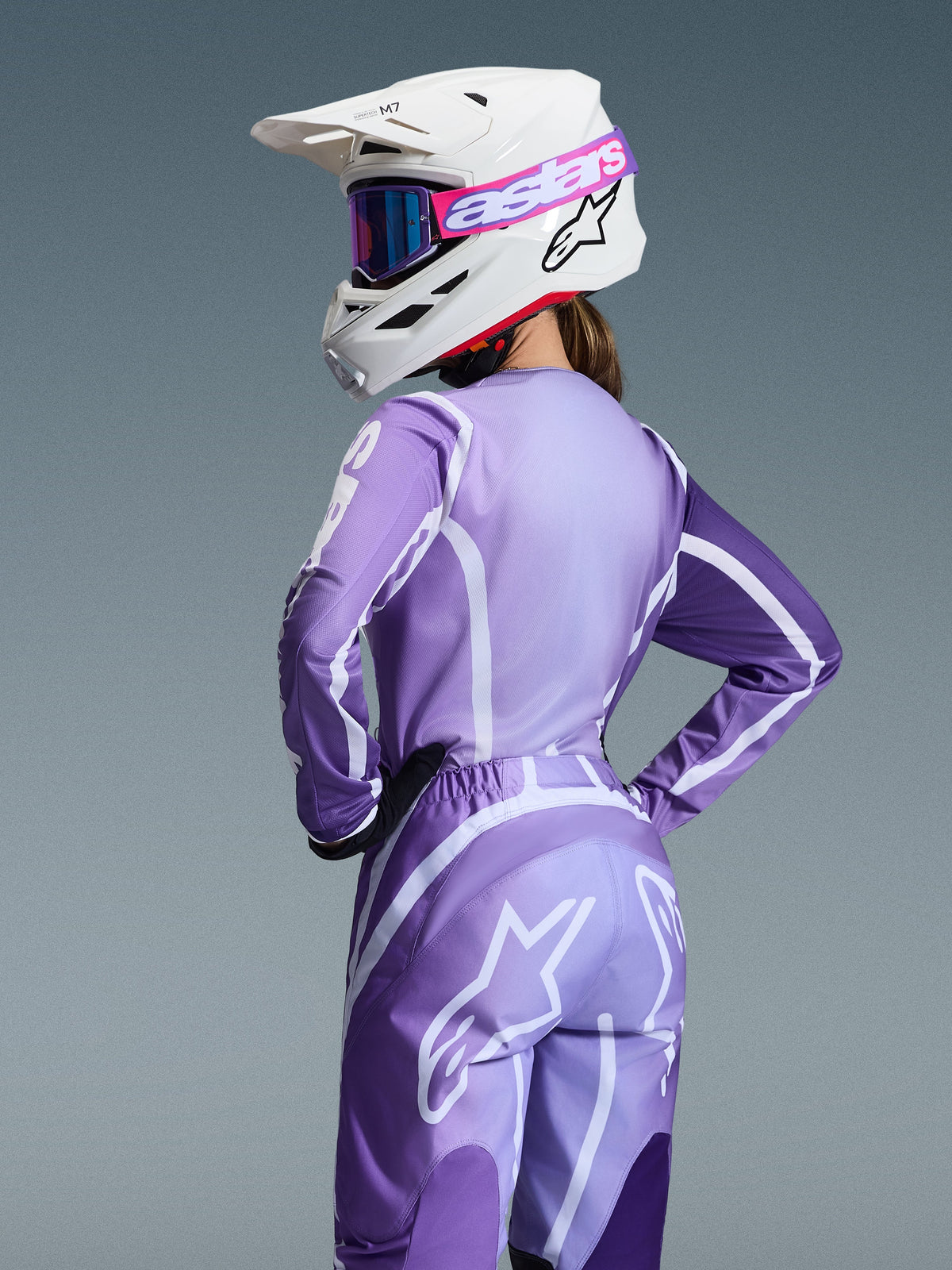 Piloto de motocross con el conjunto Alpinestars 2026 Stella Fluid Apex, jersey y pantalones púrpura y lavanda con gráficos blancos, casco blanco con gafas rosas y púrpuras, ajuste específico para mujer, equipo coordinado para carreras off-road