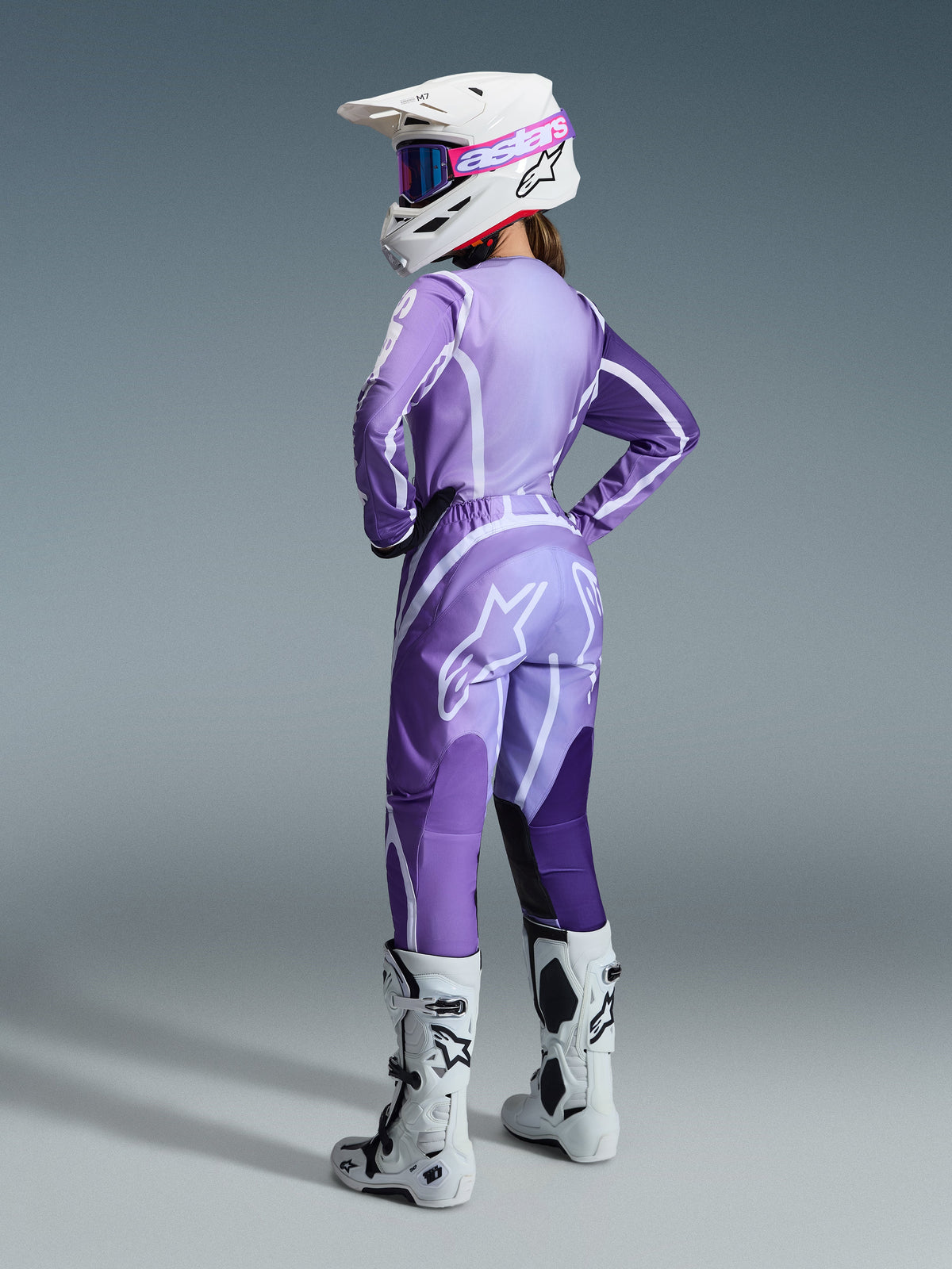 Piloto femenina de motocross con el conjunto de equipamiento Stella Fluid Apex para mujer 2026, jersey y pantalones púrpura y lavanda con detalles en blanco, casco blanco con gafas rosas, botas blancas y negras, conjunto coordinado para carreras todoterreno.