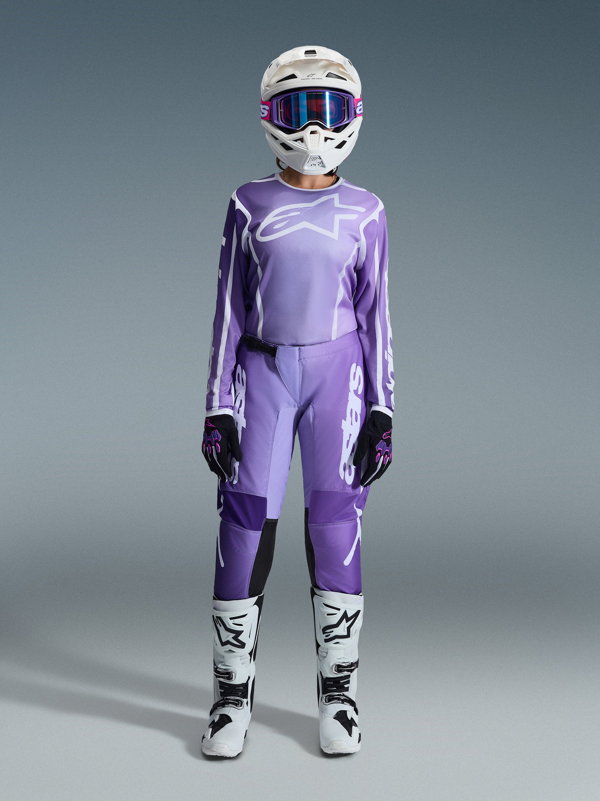 Piloto de motocross con el conjunto de equipo Alpinestars 2026 Stella Fluid Apex, jersey morado con logo blanco, pantalones morados a juego con marca en blanco, casco blanco con gafas rosas, botas blancas y negras, atuendo de motocross para mujer coordinado para carreras todoterreno