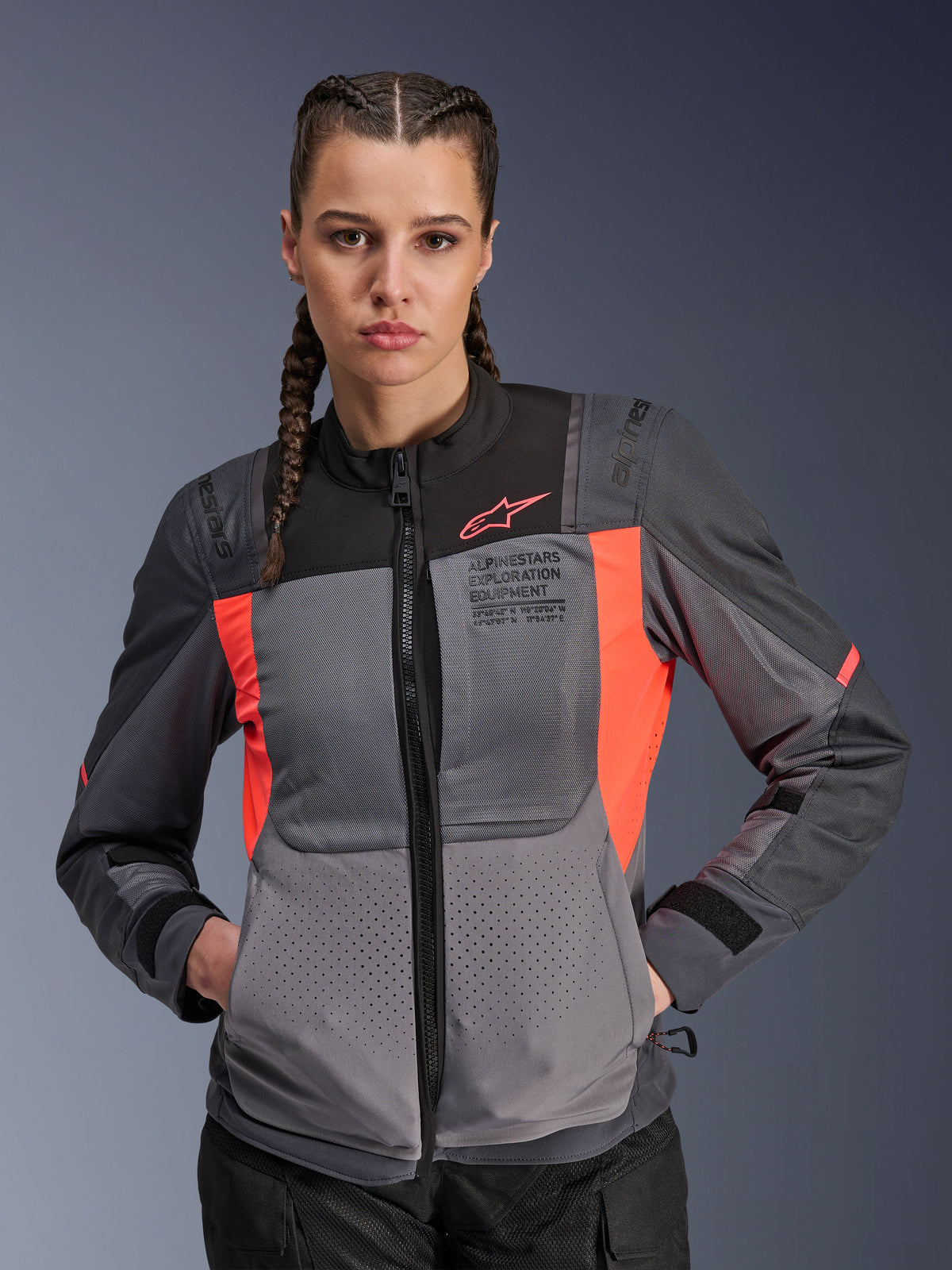 Chaqueta Alpinestars Stella ST-2 Air, chaqueta de moto para mujer, gris oscuro y coral flúor, usada por una modelo con el pelo trenzado, con paneles de malla técnica para el flujo de aire, perforaciones láser y marca 'Alpinestars Exploration Equipment' en el pecho, equipamiento de touring para verano