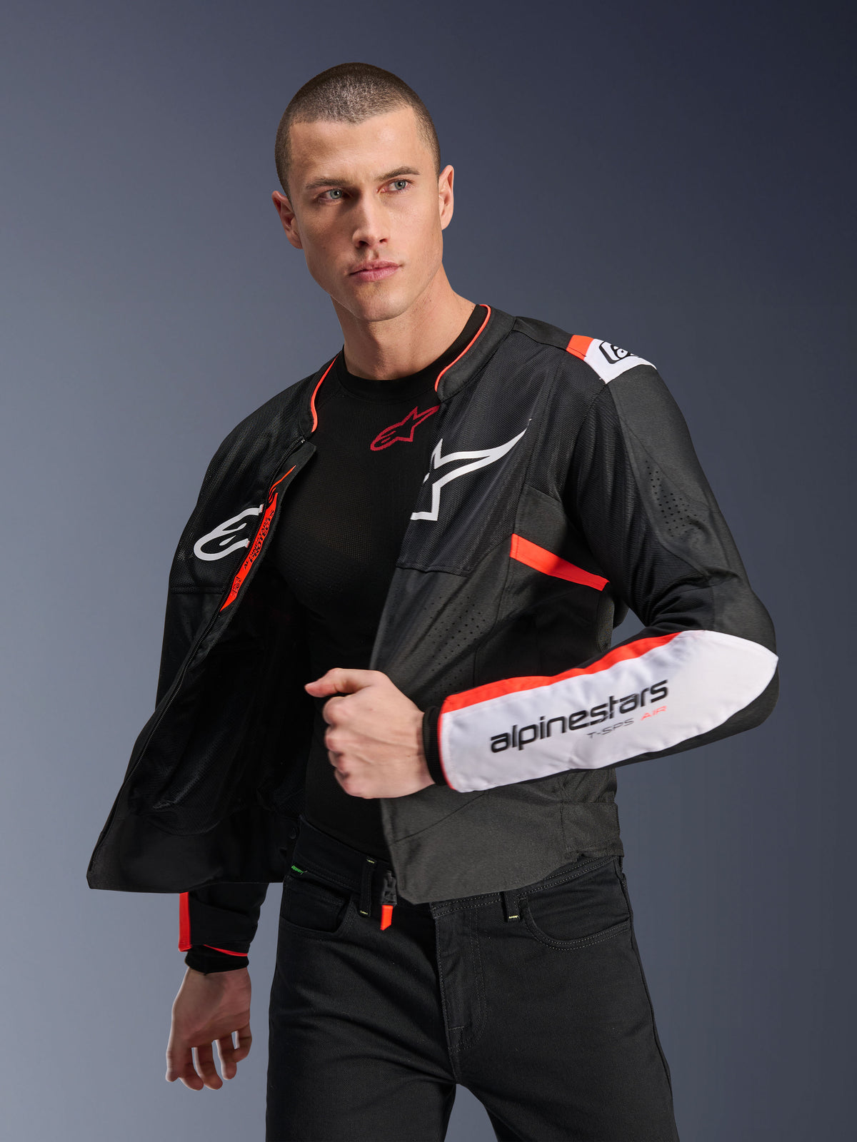 Modelo masculino con la chaqueta Alpinestars T-SPS Air V2, chaqueta textil de moto en color negro con detalles en blanco y rojo fluorescente, paneles de malla para ventilación, logotipo blanco de Alpinestars en el pecho y marca en la manga, usada abierta sobre una prenda base negra