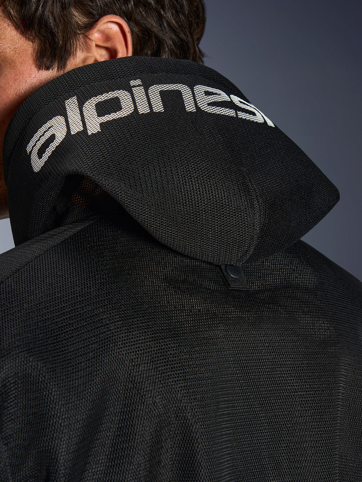 Alpinestars Chrome Superairflow Hoodie, negra, vista de detalle de la capucha de malla 3D con logotipo de Alpinestars en blanco, botón de presión de estabilización trasero, equipamiento textil ventilado para moto en condiciones de calor extremo