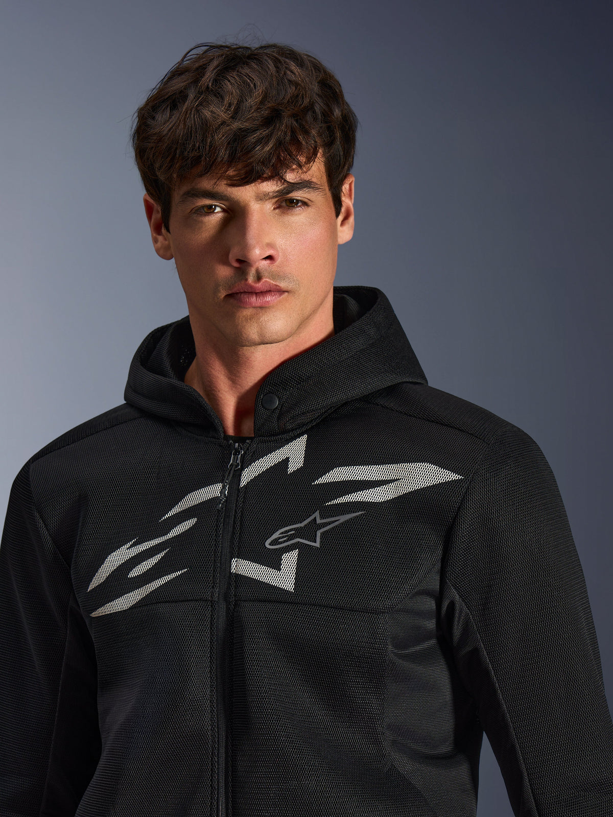 Alpinestars Chrome Superairflow Hoodie, sudadera de moto negra, cuenta con construcción de malla 3D con un gran logotipo gráfico blanco en el pecho, lucida por un modelo, prenda de conducción deportiva urbana con un moderno diseño ventilado