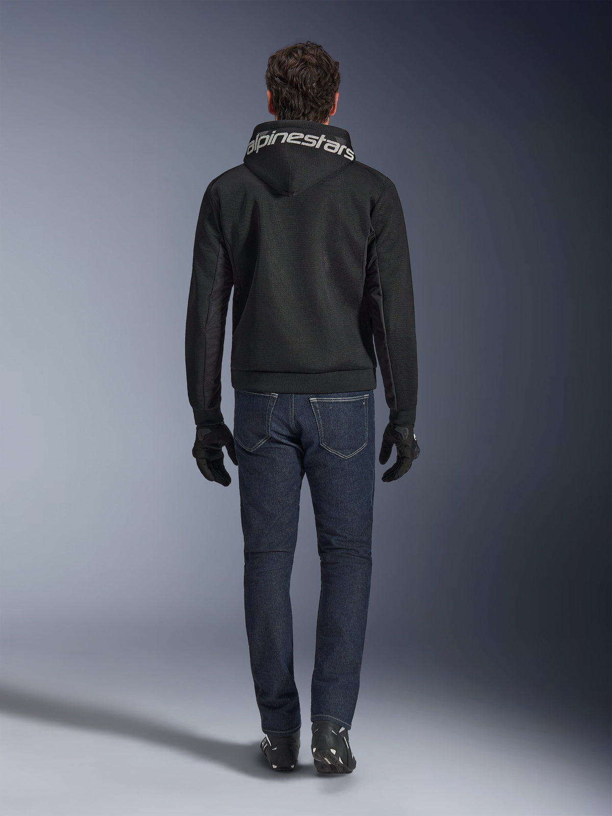 Sudadera con capucha Alpinestars Chrome Superairflow, sudadera de moto negra con logotipo blanco en la capucha, vista desde atrás, combinada con vaqueros azul oscuro, guantes negros y botas blancas y negras, equipamiento de conducción urbana y deportiva