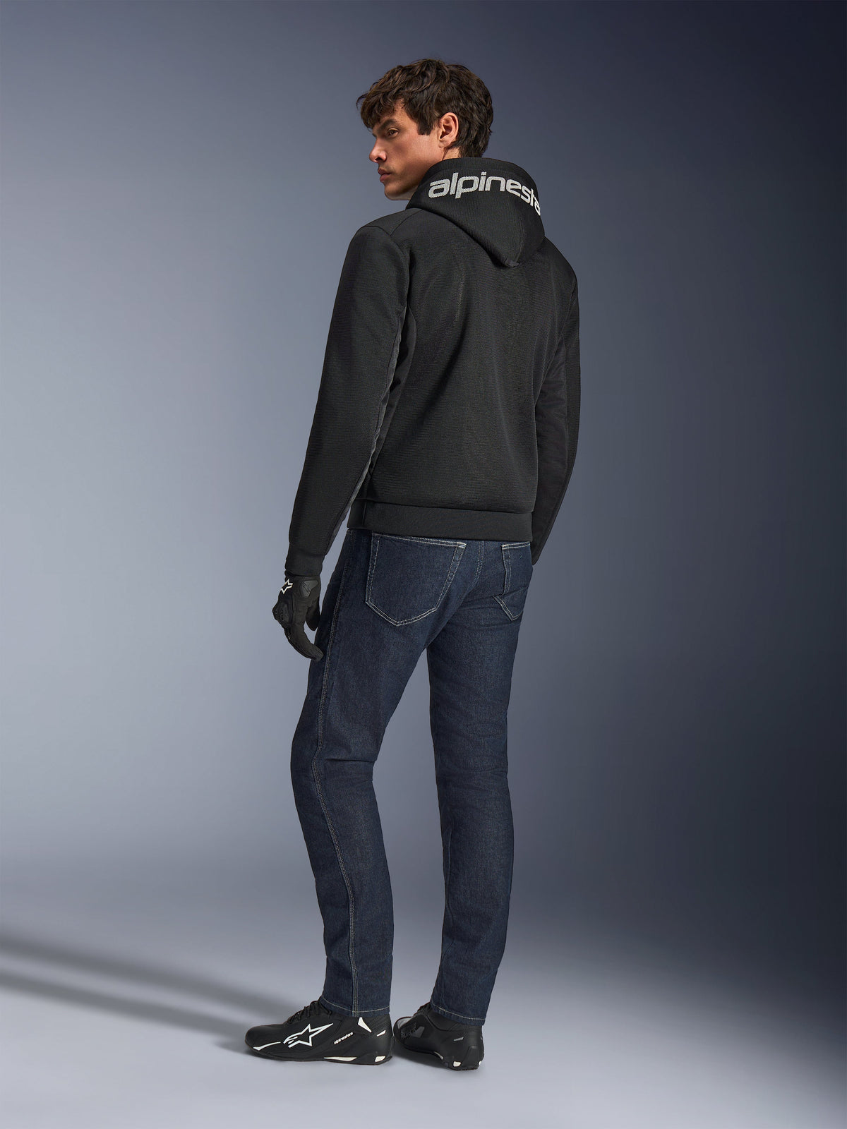 Sudadera con capucha Alpinestars Chrome Superairflow, negra, vista posterior en modelo masculino, con la marca Alpinestars en blanco en la capucha, combinada con vaqueros denim oscuros y guantes negros, diseño de equipamiento urbano para motociclismo