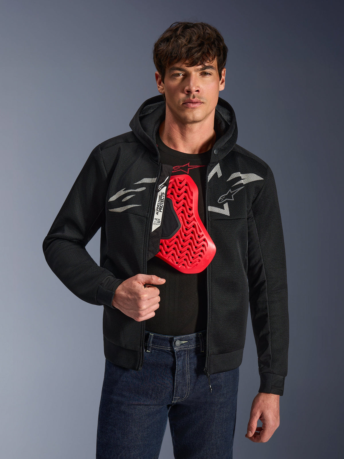 Alpinestars Chrome Superairflow Hoodie en negro, con logotipos Astars en blanco y gris, usada por un modelo masculino con un protector de pecho rojo visible debajo, tejido de malla 3D para ventilación, ropa de moto urbana deportiva.