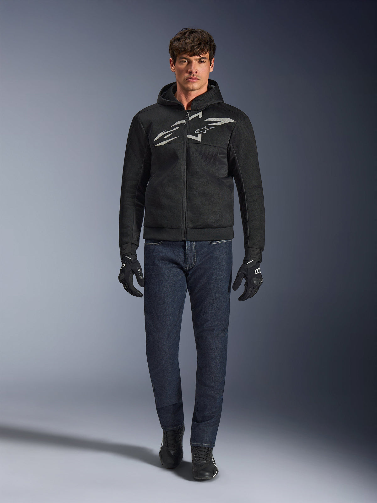 Motociclista con la sudadera con capucha Alpinestars Chrome Superairflow en negro y gris, combinada con vaqueros azul oscuro, guantes de moto negros y calzado negro. La sudadera presenta un gran logotipo estilizado en el pecho y una textura de malla 3D para un alto rendimiento en temperaturas elevadas.
