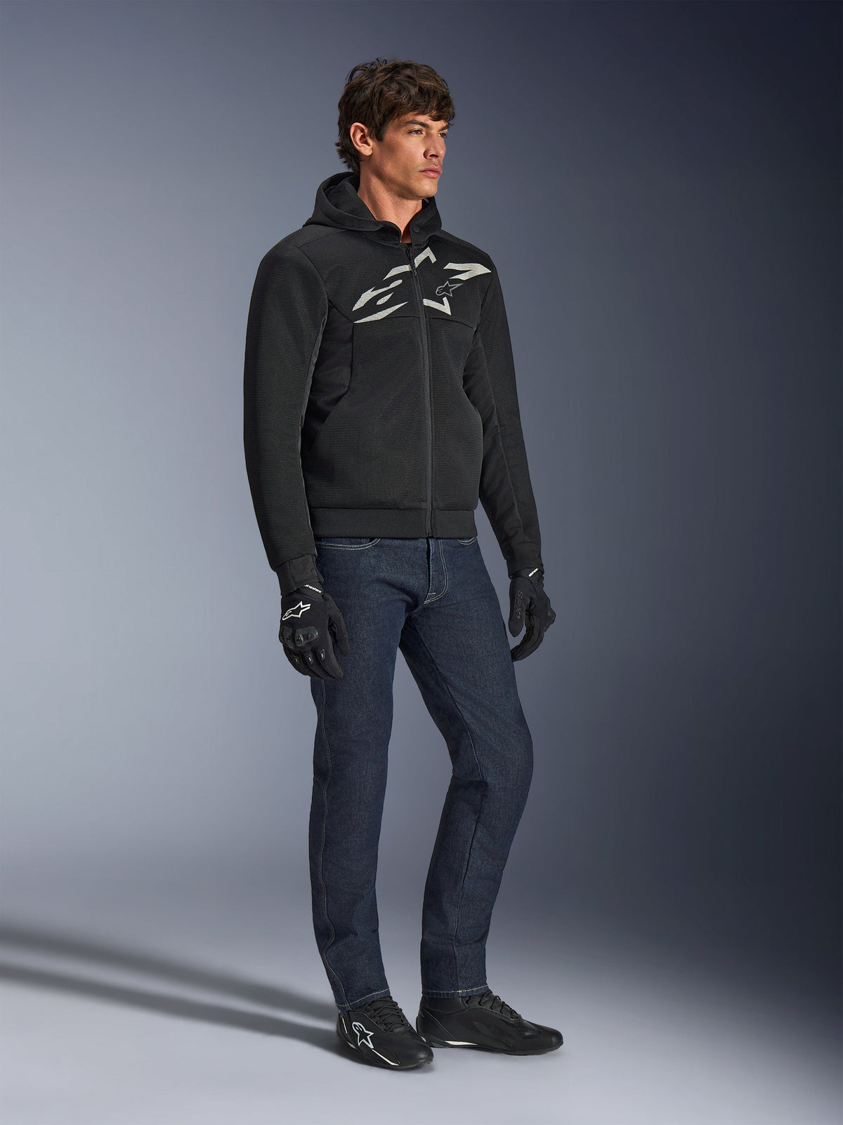 Sudadera con capucha Alpinestars Chrome Superairflow, negra con logotipo en blanco y gris, construcción textil de malla 3D ventilada, combinada con vaqueros denim azul oscuro y guantes de moto negros, estilo de conducción deportiva urbana.