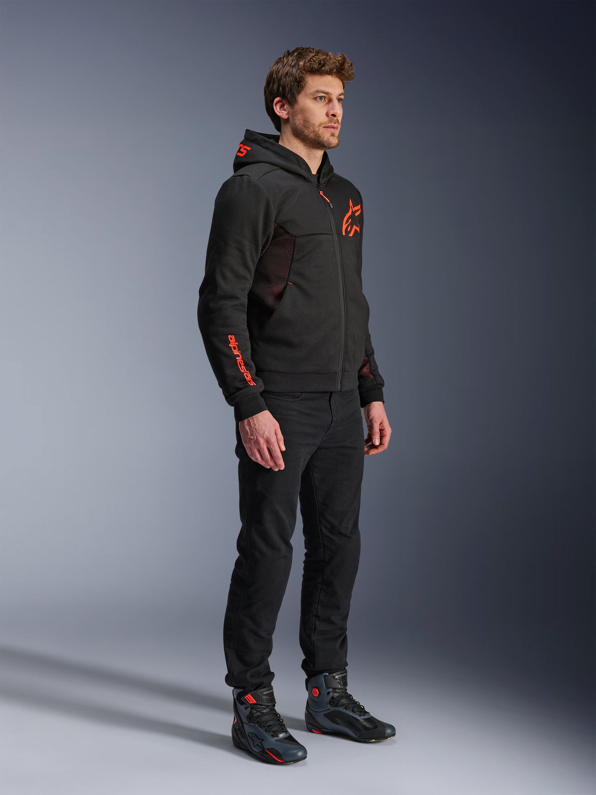 Alpinestars Chrome Air Sport Hoodie, Negro Rojo Flúor, sudadera de conducción urbana con forro polar técnico, combinada con pantalones negros y botas de moto, con paneles de ventilación de malla y logotipos en rojo flúor en el pecho y la manga