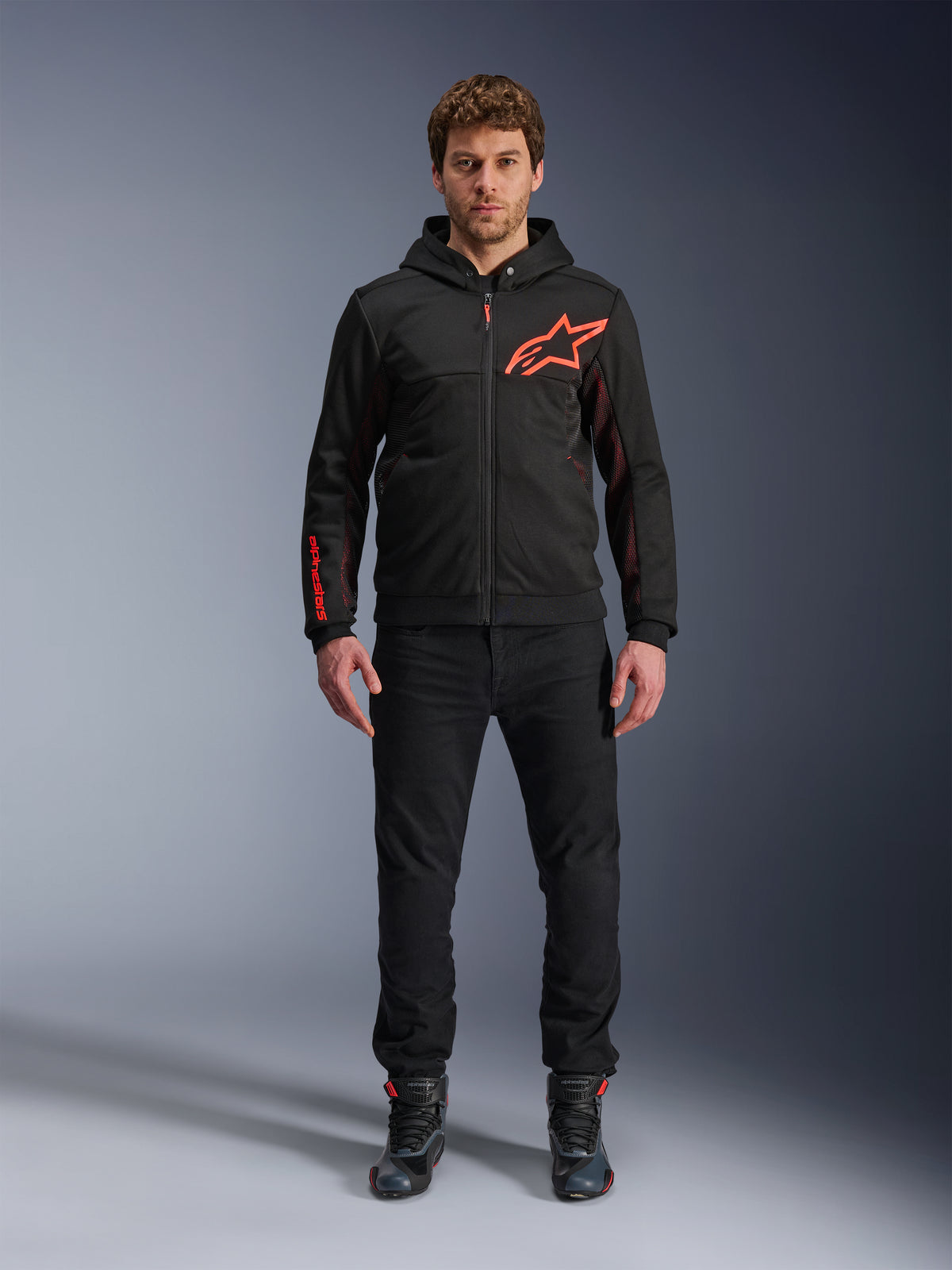 Sudadera Alpinestars Chrome Air Sport, negro y rojo flúor, chaqueta urbana para motociclista con construcción de tejido polar técnico, con un gran logotipo rojo de Astars en el pecho y marca en la manga, lucida por un modelo con pantalones negros y calzado de moto