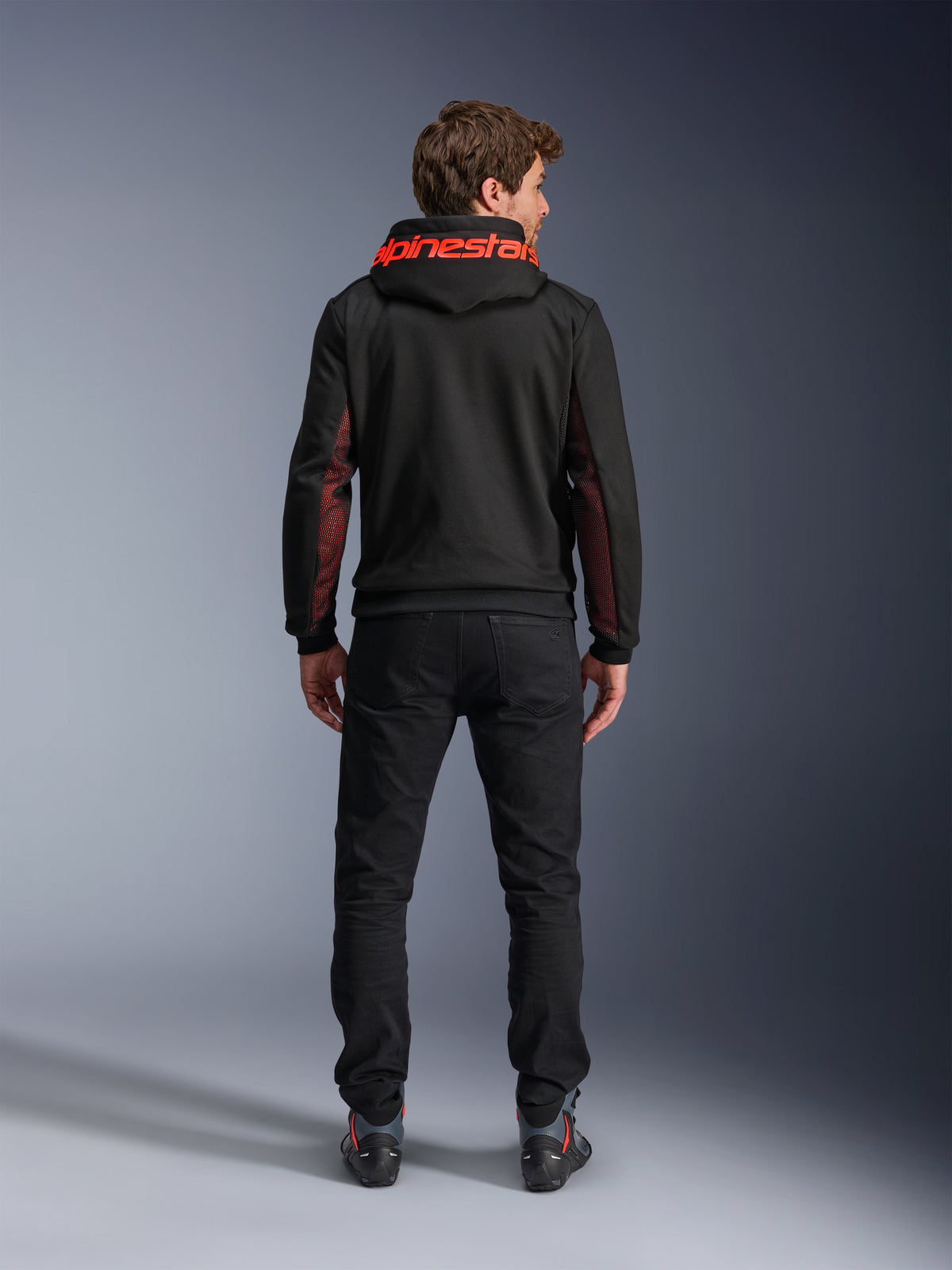 Alpinestars Chrome Air Sport Hoodie, negro y rojo fluo, vista posterior de un hombre con chaqueta de moto casual, con la marca Alpinestars en rojo en la capucha y paneles de malla rojos en las mangas, combinada con pantalones negros.