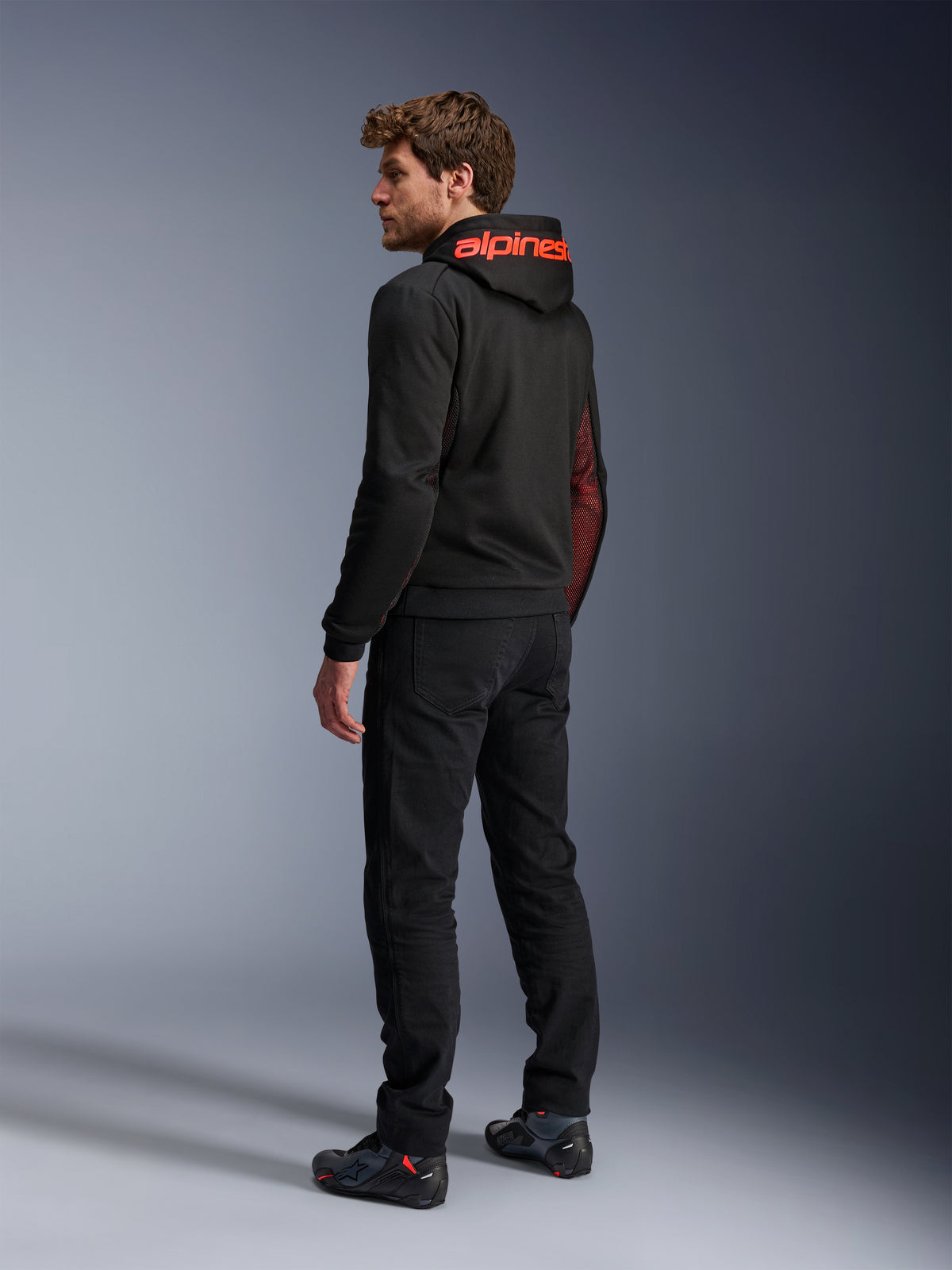 Sudadera Alpinestars Chrome Air Sport, sudadera de moto urbana, negro y rojo flúor, vista trasera que muestra la marca Alpinestars en rojo en la capucha y los paneles laterales de malla, usada con pantalones de moto negros y calzado técnico