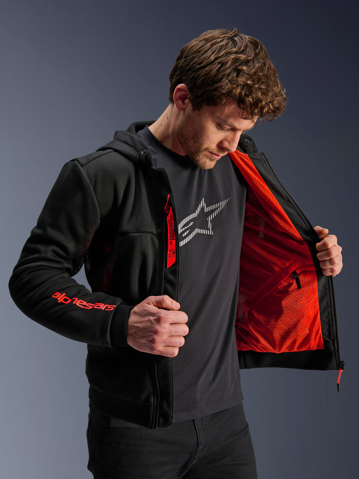 Alpinestars Chrome Air Sport Hoodie en Negro y Rojo Flúor, modelo masculino abriendo la chaqueta para mostrar el forro de malla rojo brillante, logotipo Alpinestars en la manga derecha y cremallera del pecho, chaqueta de moto urbana casual diseñada para el flujo de aire.