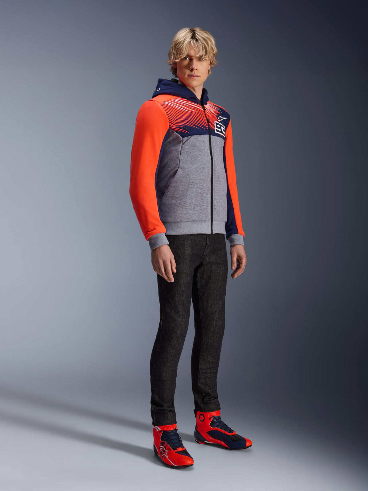 Alpinestars MM93 Chrome V2 Sport Hoodie, urban motorcycle hoodie, gray melange body with navy blue chest and bright red fluo sleeves, featuring the 93 logo on the chest, worn by a model with slim-fit black pants and coordinated red and blue high-top riding shoes, modern athletic fit for street riding