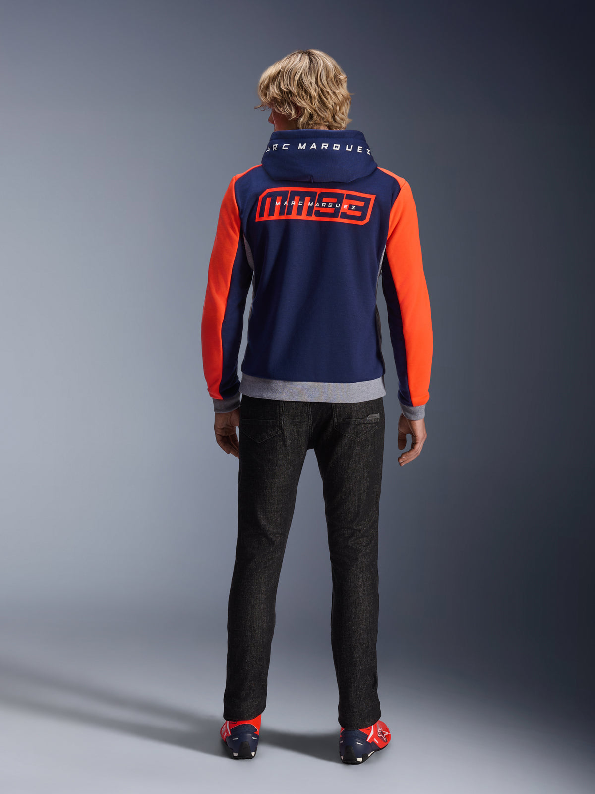 Model from the back wearing the Alpinestars MM93 Chrome V2 Sport Hoodie, Blue Navy with Bright Red Fluo sleeves and Gray Mel details, large MM93 and Marc Marquez logos on the back and hood, casual motorcycle riding hoodie paired with black pants