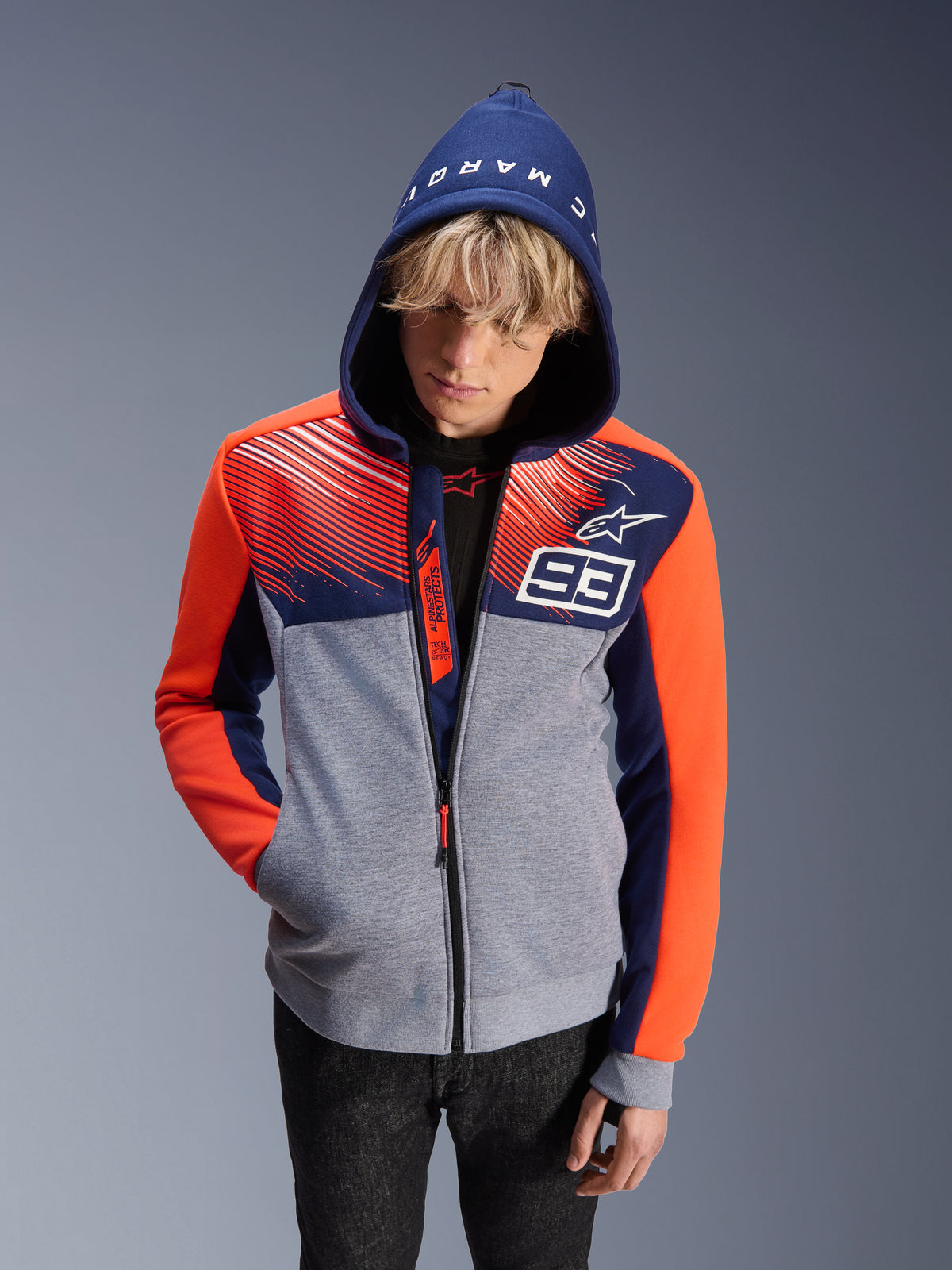 Alpinestars MM93 Chrome V2 Sport Hoodie, motorcycle riding hoodie, gray melange body with navy blue upper and fluorescent orange sleeves, featuring white number 93 and Alpinestars star logo, Marc Marquez text on the hood, worn by a male model with hood up, urban motorsport-inspired design